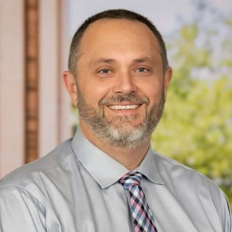 Portrait of a smiling man with a trimmed beard, short hair, wearing a light gray dress shirt and a pink, gray, and black plaid tie, standing outdoors with blurred green trees in the background.