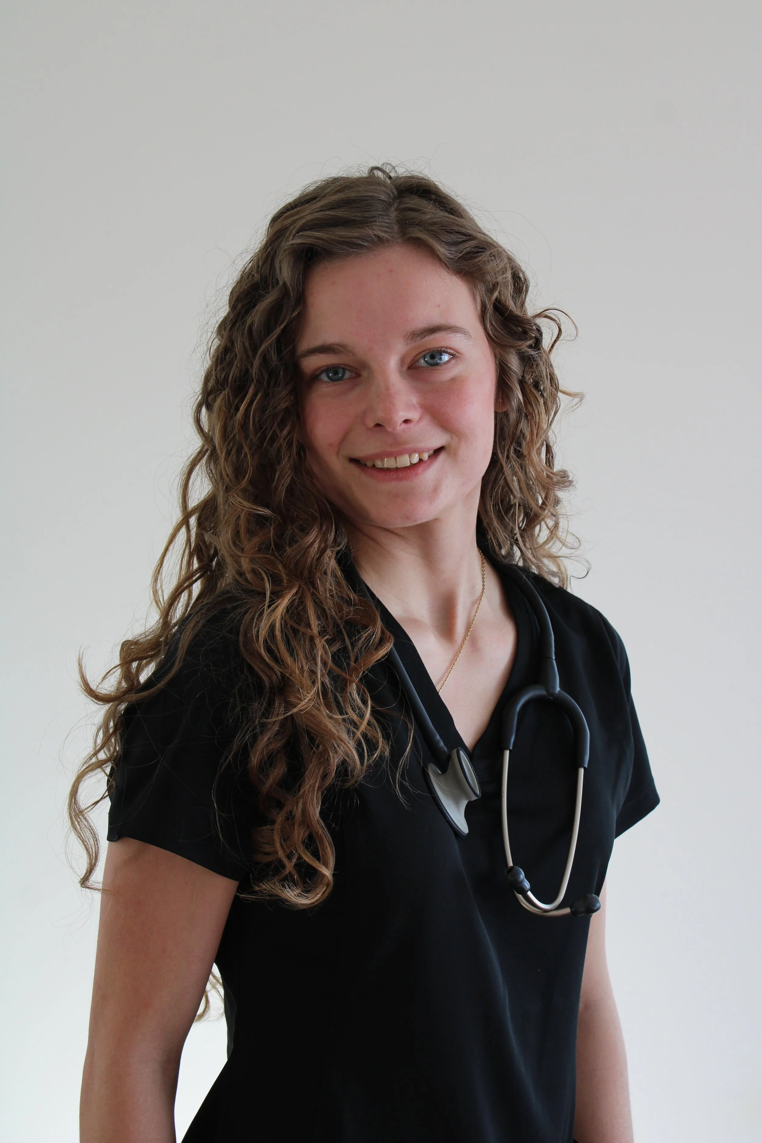 A young woman with long, curly brown hair and blue eyes, wearing black medical scrubs and a stethoscope around her neck, smiling at the camera against a plain white background.
