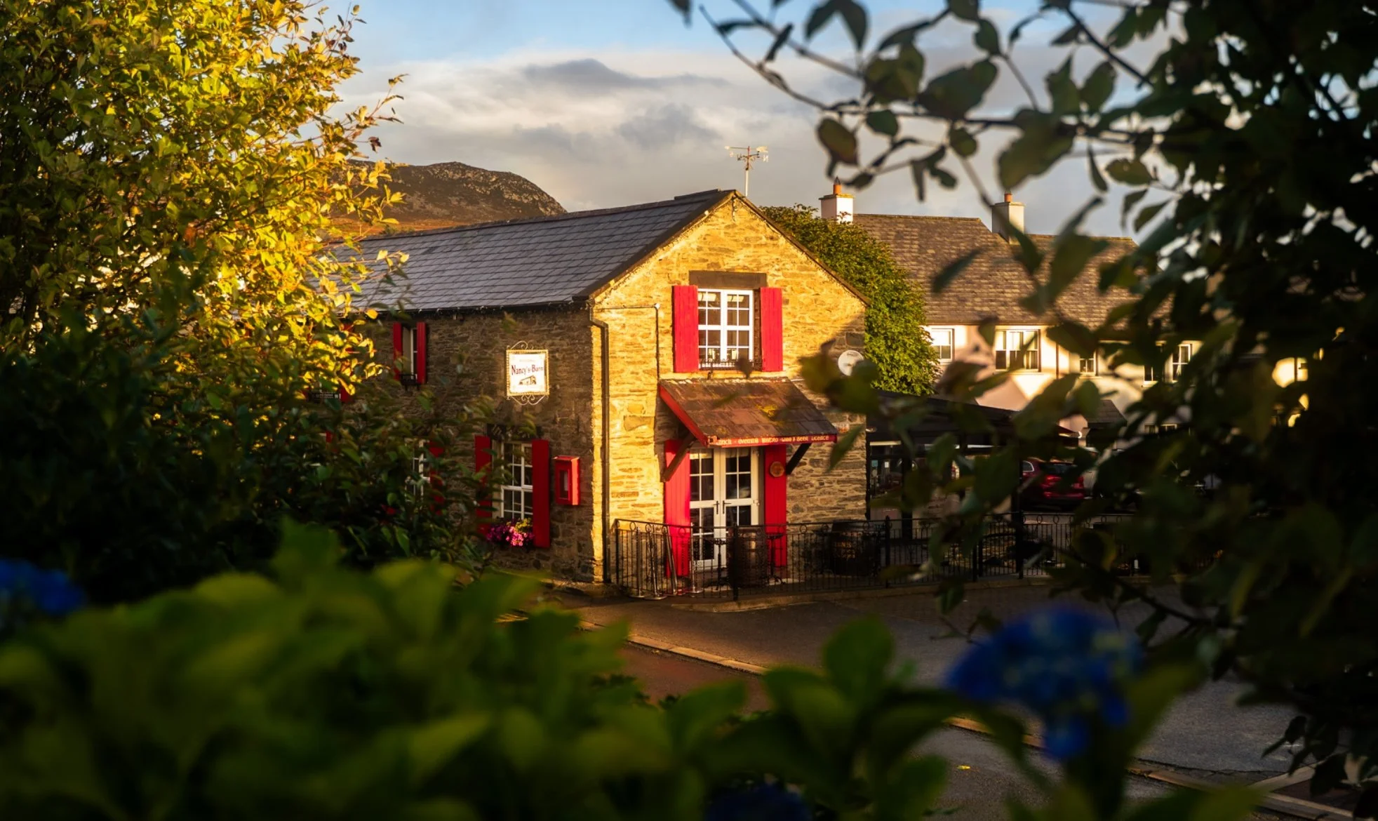 Nancys Barn, a stone building with red window shutters, surrounded by green foliage, during sunset.