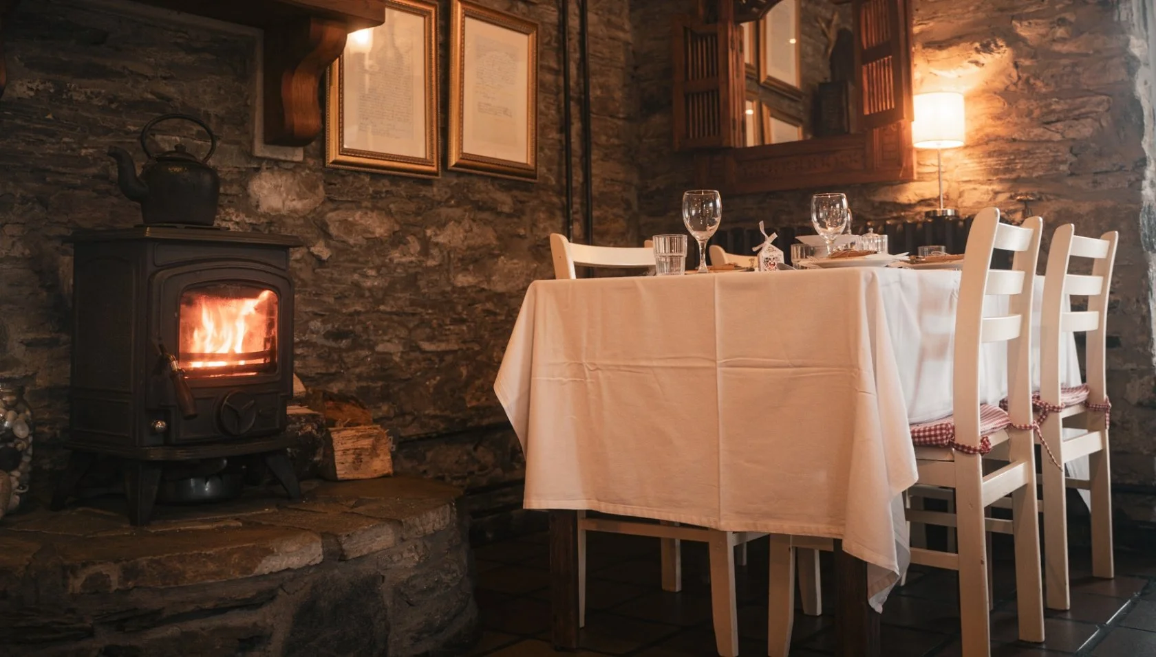 A cozy dining room with a stone fireplace, a wooden table covered with a white tablecloth, and white chairs with red and white checkered cushions. The table is set with wine glasses, plates, and cutlery. Warm lighting comes from a lamp and the fireplace.