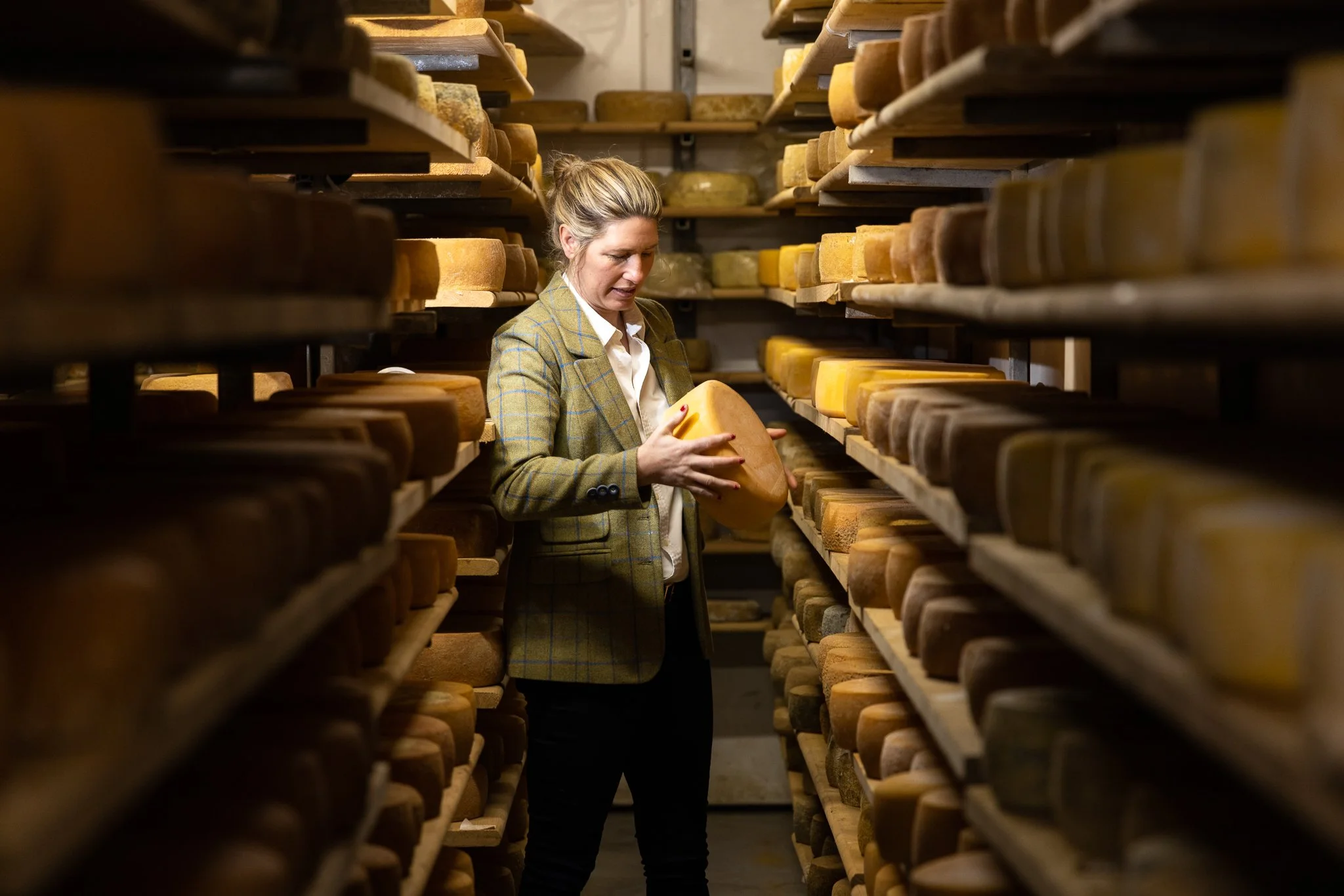 A woman in a plaid blazer examining a large wheel of cheese in a cheese aging cellar with shelves of similar cheese wheels surrounding her.