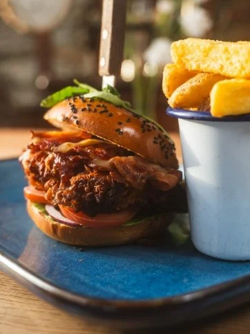 Close-up of a chicken burger with, lettuce, tomato, and a seeded bun, served with chips in a white container, on a blue plate.