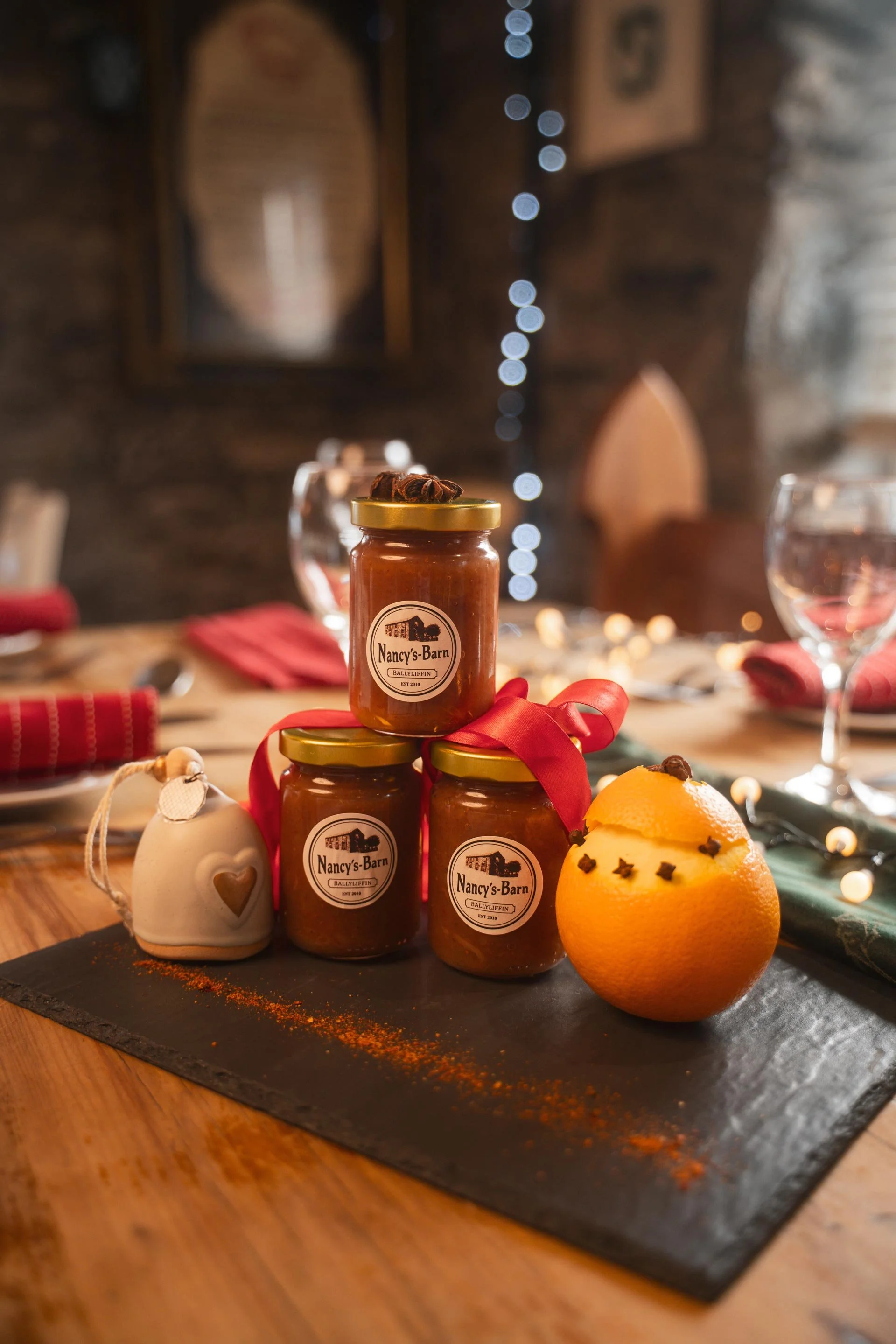 A display of three small jars labeled Nancy's Barn on a round black slate with an orange, star-shaped fruit with cloves inserted for decoration. The jars are arranged in a pyramid with a red ribbon tied around the middle jar. A small white ceramic heart ornament and a white bauble with a heart cutout are also on the slate. Blurred background shows a table setting with glasses, napkins, and decorative string lights in a cozy, rustic room.