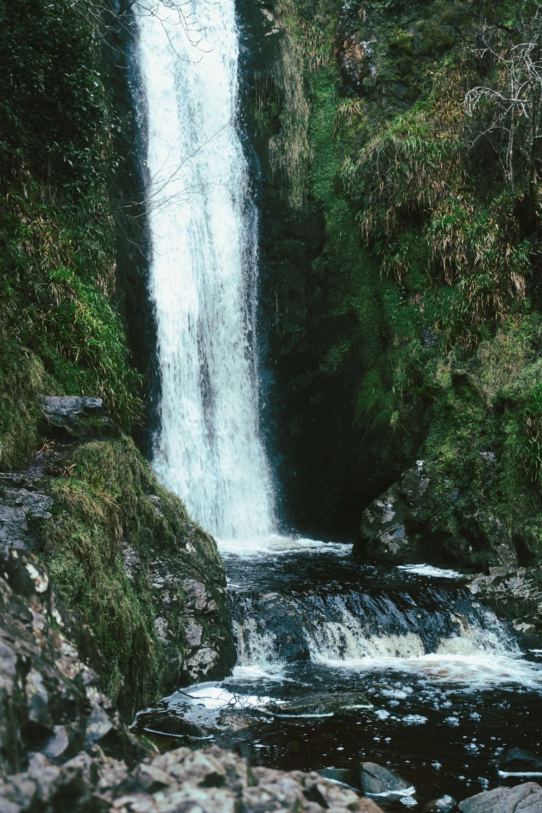 Glenevin waterfall flowing into a rocky stream surrounded by green foliage.