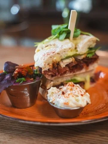 A sandwich with layers of meat, lettuce, and bread, served with two side dishes in small bowls on a wooden plate.