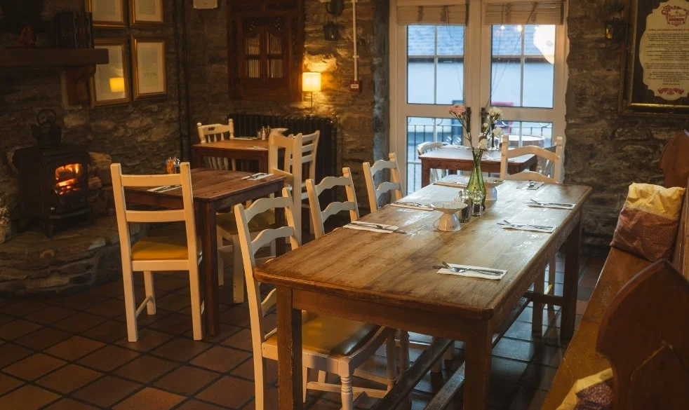 Cozy restaurant with a wooden table set for dining, decorated with a vase of flowers, and surrounded by white chairs, with a stone wall and a fireplace in the background.