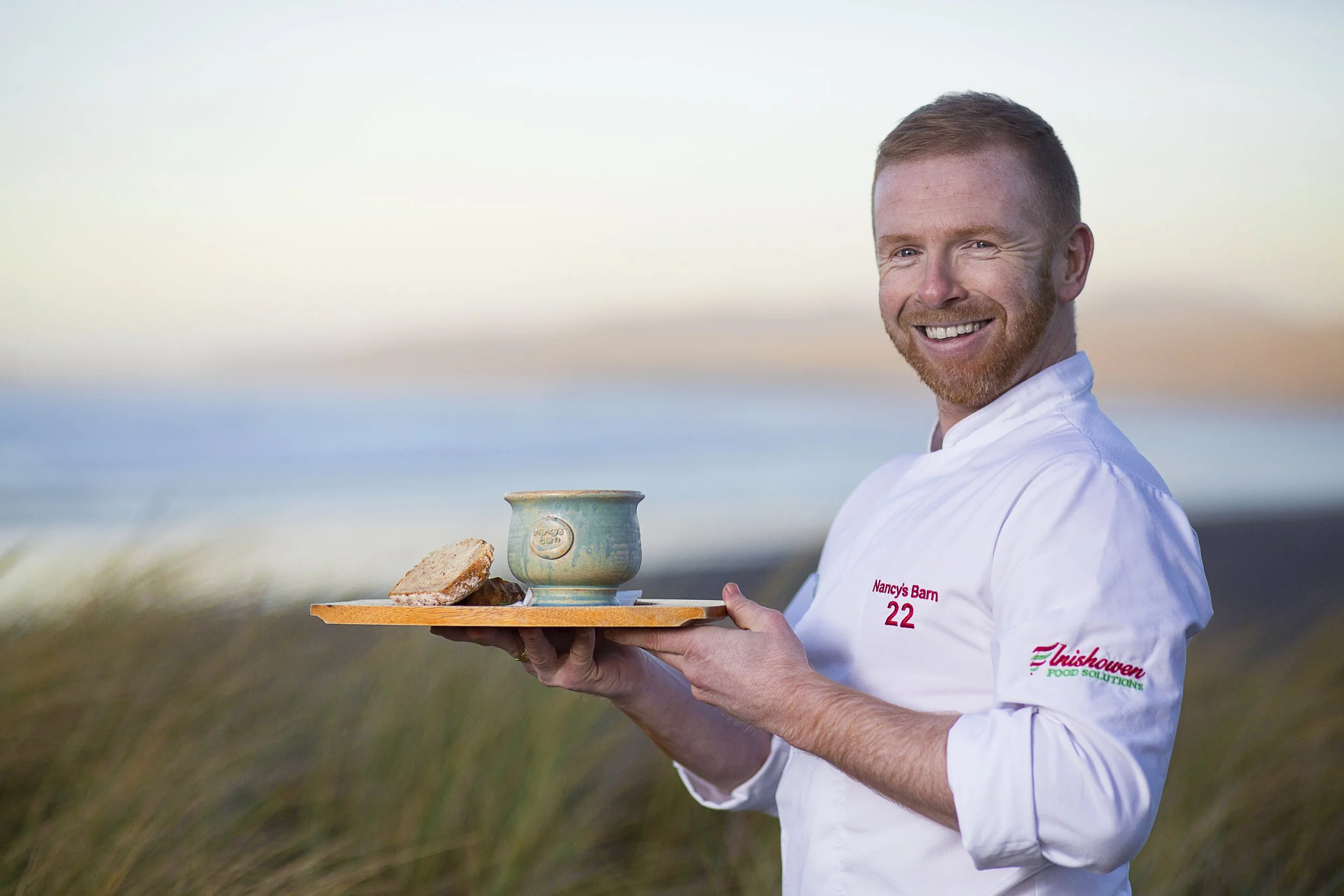 Chef Kieran Doherty holding a handmade Moville Pottery chowder bowl on a tray by the sea