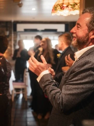 Man clapping at a social gathering in a warmly lit room with other people in the background.
