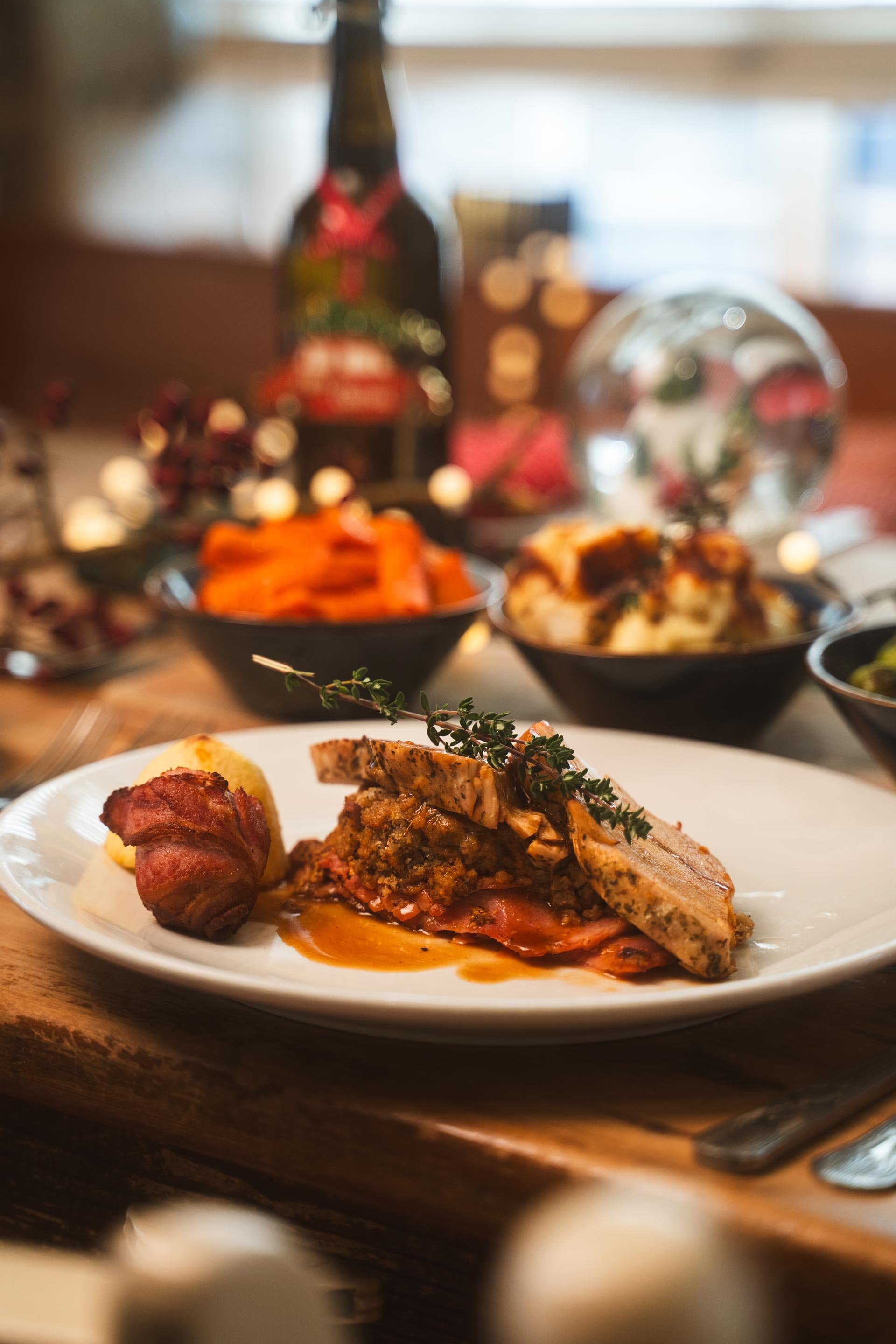 Close-up of a plate of served food including bacon-wrapped meat with sauce, stuffing, and mashed potatoes with gravy at a festive dinner setting.