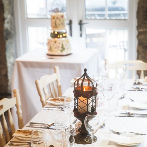 Dinner table set with white tablecloth, wine glasses, silverware, and napkins, with a black lantern centerpiece, and a wedding cake on a table in the background.