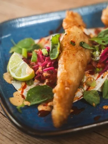 Fried fish tacos on a bed of salad with lime wedges and various greens, sauce, and pink pickled vegetables on a blue plate.