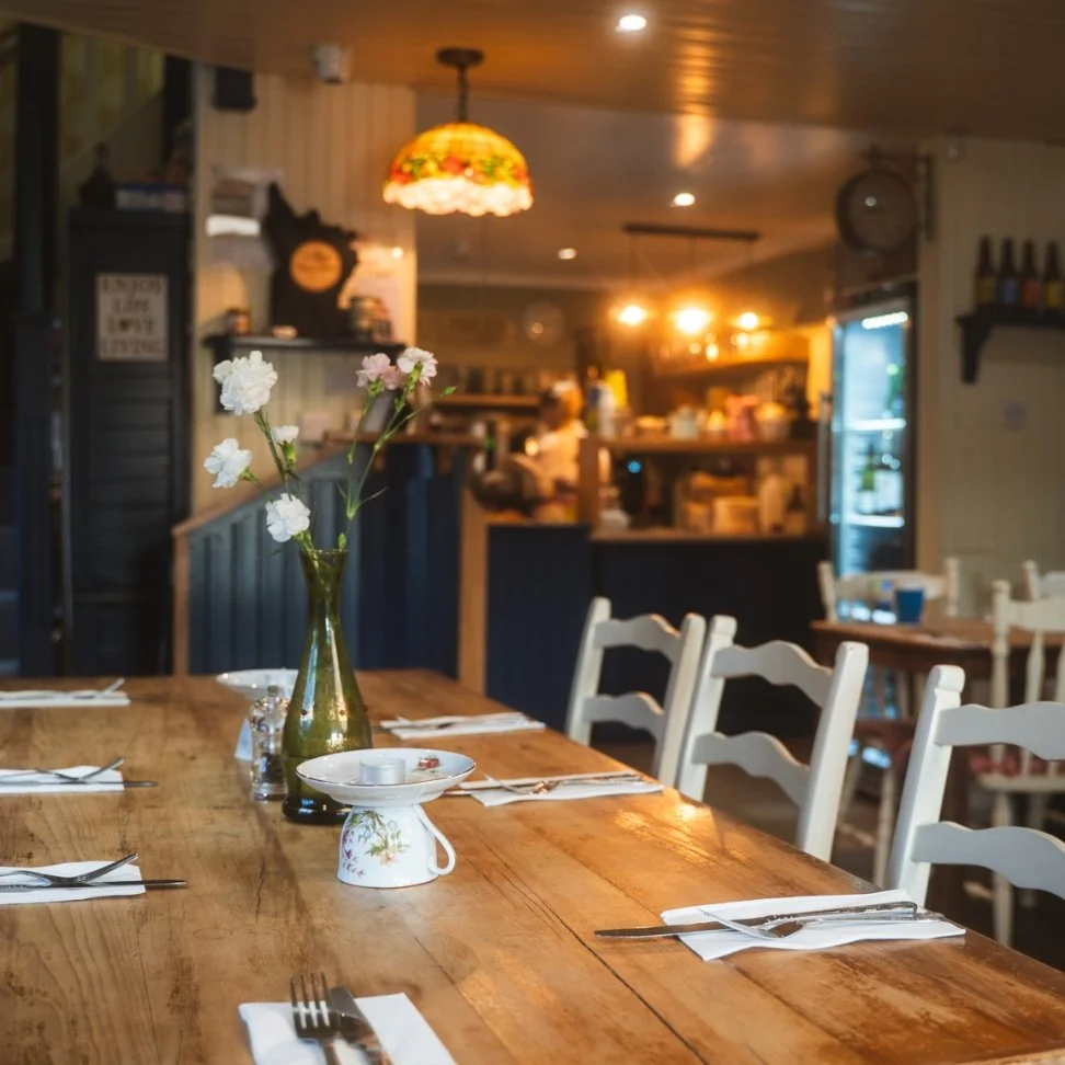 Nancy's cozy restaurant interior with a large wooden table set with utensils and napkins, a vase with white flowers as a centerpiece, warm lighting, and a visible kitchen in the background.