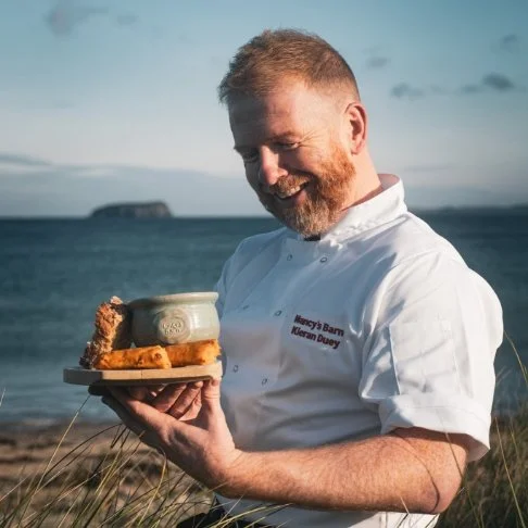Kieran Doherty holding a bowl of seafood chowder, outdoors near the ocean with Glashedy island in the background.