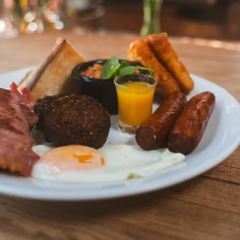 A breakfast plate with toast, fried egg, sausages, a slice of bacon, crispy hash browns, a small bowl of salsa, and a shot glass of orange juice.