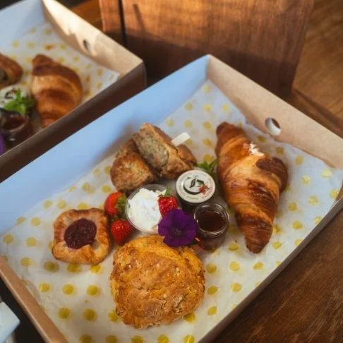 A bakery-style breakfast basket with a croissant, muffin, fruit tart, fried chicken strip, whipped cream, strawberries, jam, and a purple flower on a yellow polka dot paper in a takeout box.