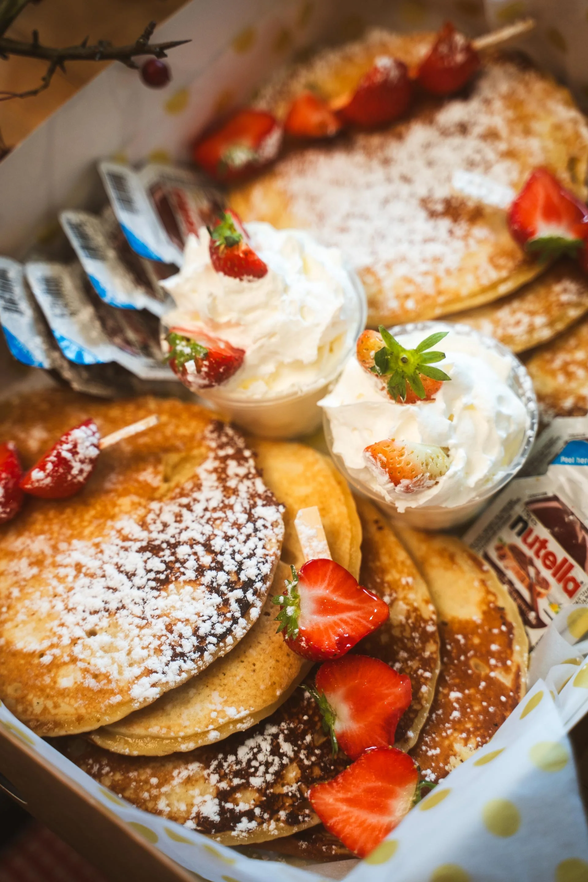 Stacked pancakes dusted with icing sugar, with fresh strawberries, whipped cream and Nutella sachets