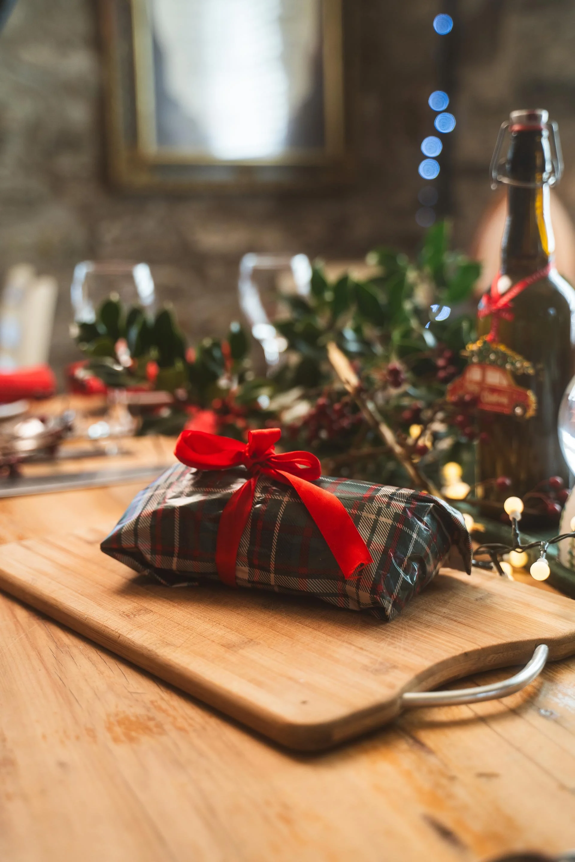 A Christmas gift wrapped in plaid paper with a red ribbon bow, placed on a wooden cutting board on a table. In the background, there is blurry holiday table decor including a bottle, green foliage, and string lights.