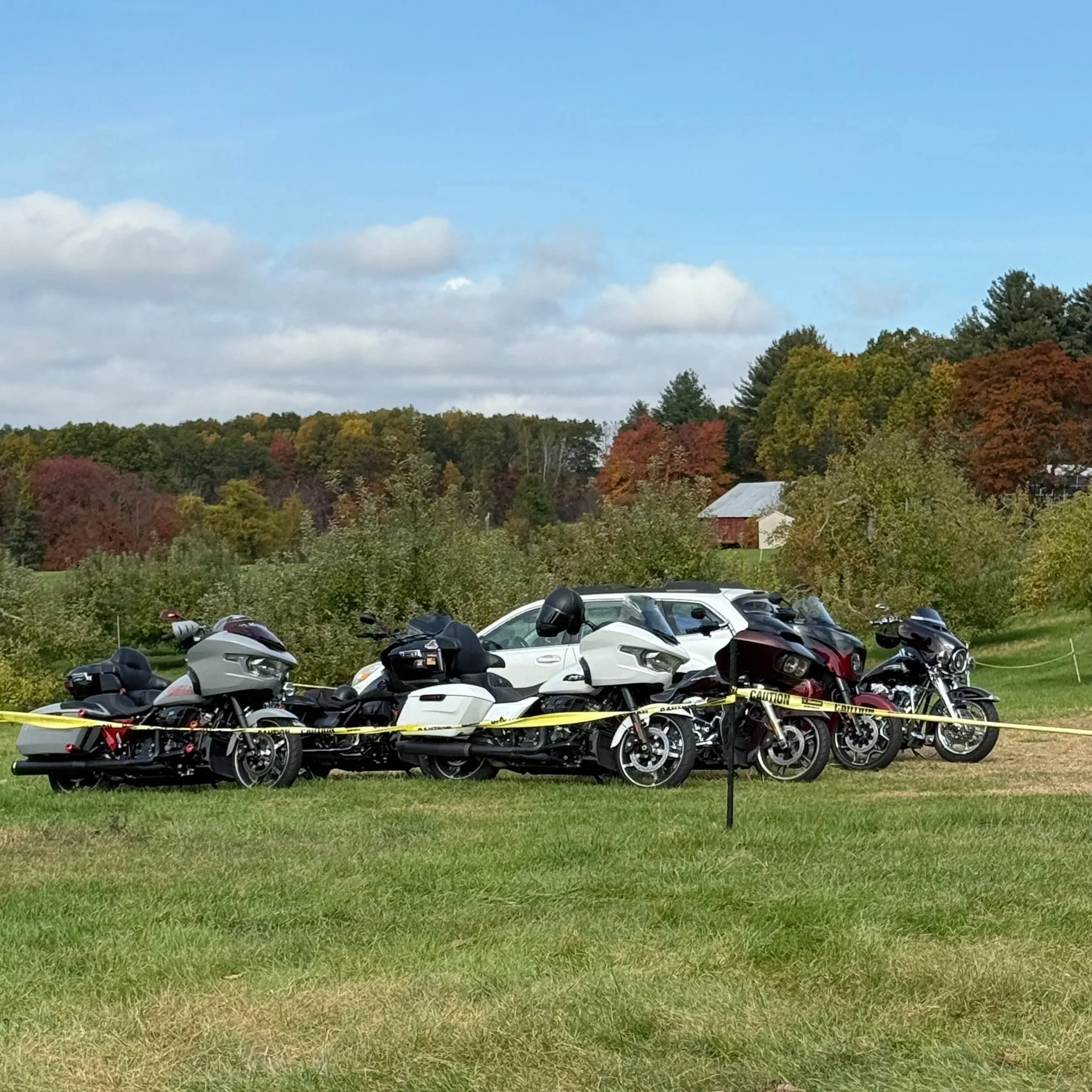 Multiple motorcycles parked on grass, surrounded by yellow caution tape, with trees and a barn in the background under a partly cloudy sky.