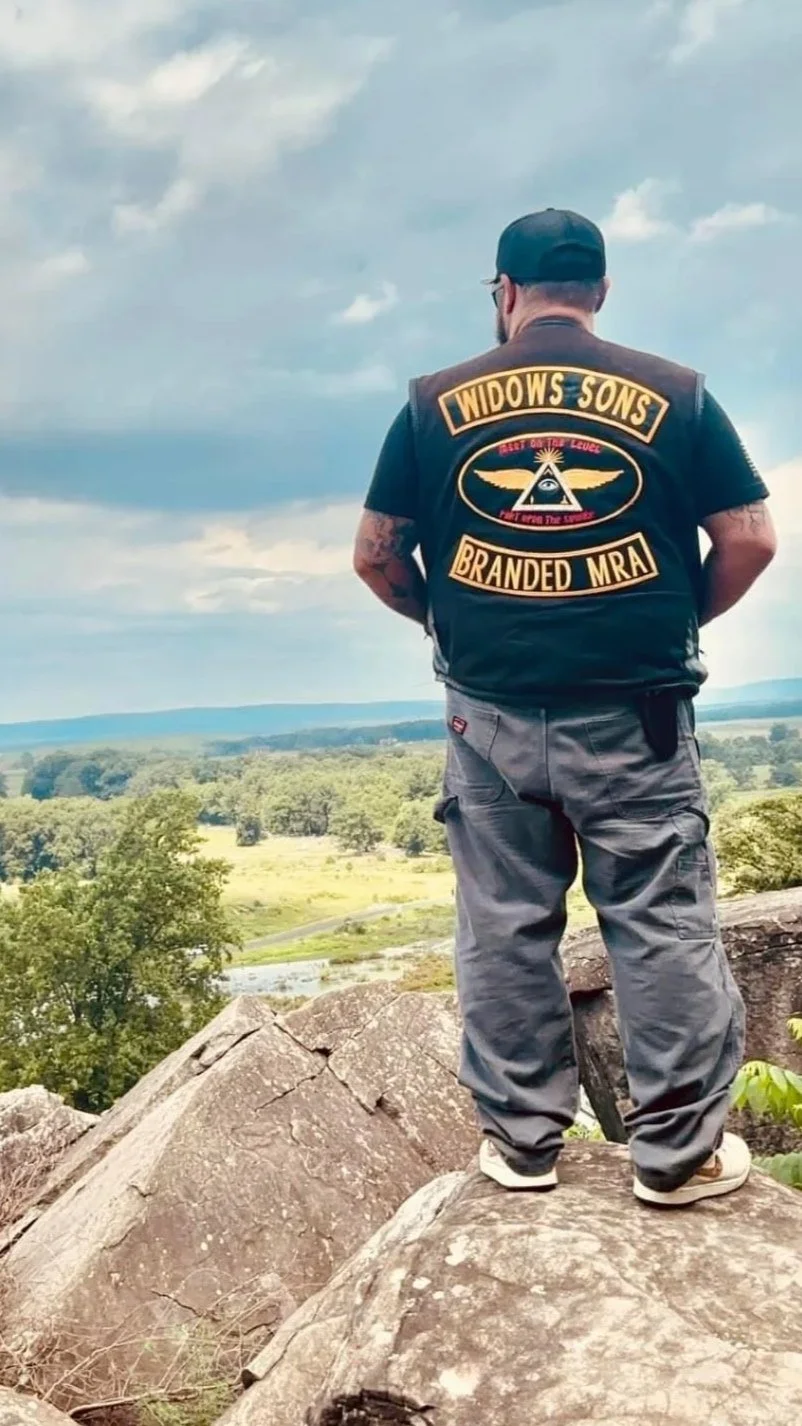 A man standing on a rock overlooking a scenic landscape with trees, water, and hills, wearing a leather vest with patches that say "Widows Sons Branded MRA" and an emblem with an all-seeing eye.