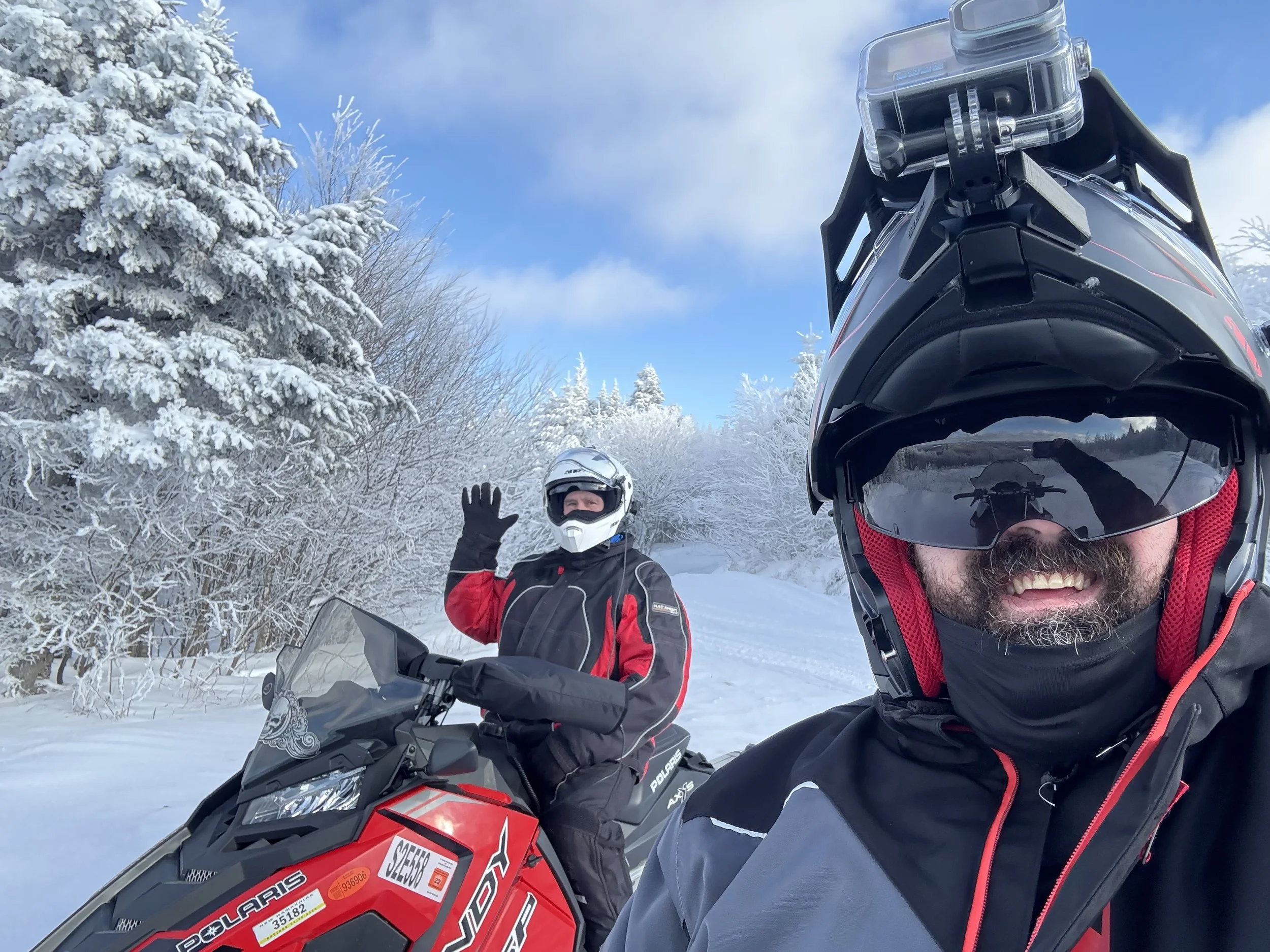 Two men wearing helmets and winter gear on snowmobiles in a snowy landscape with snow-covered trees and a partly cloudy blue sky, one man smiling and the other waving.