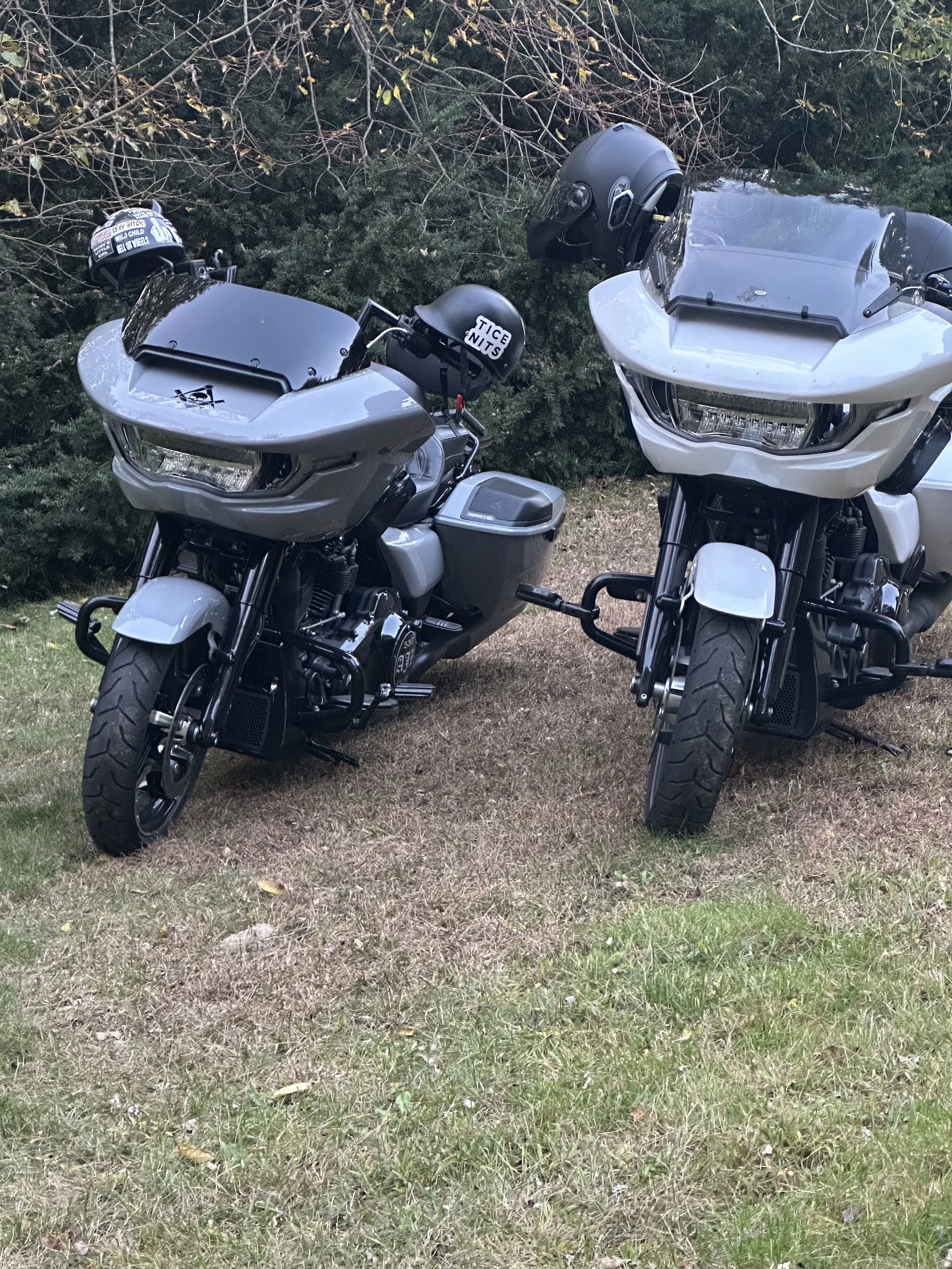 Two grey touring motorcycles parked on grass with trees in the background, both helmets hanging on the handlebars, one with stickers.