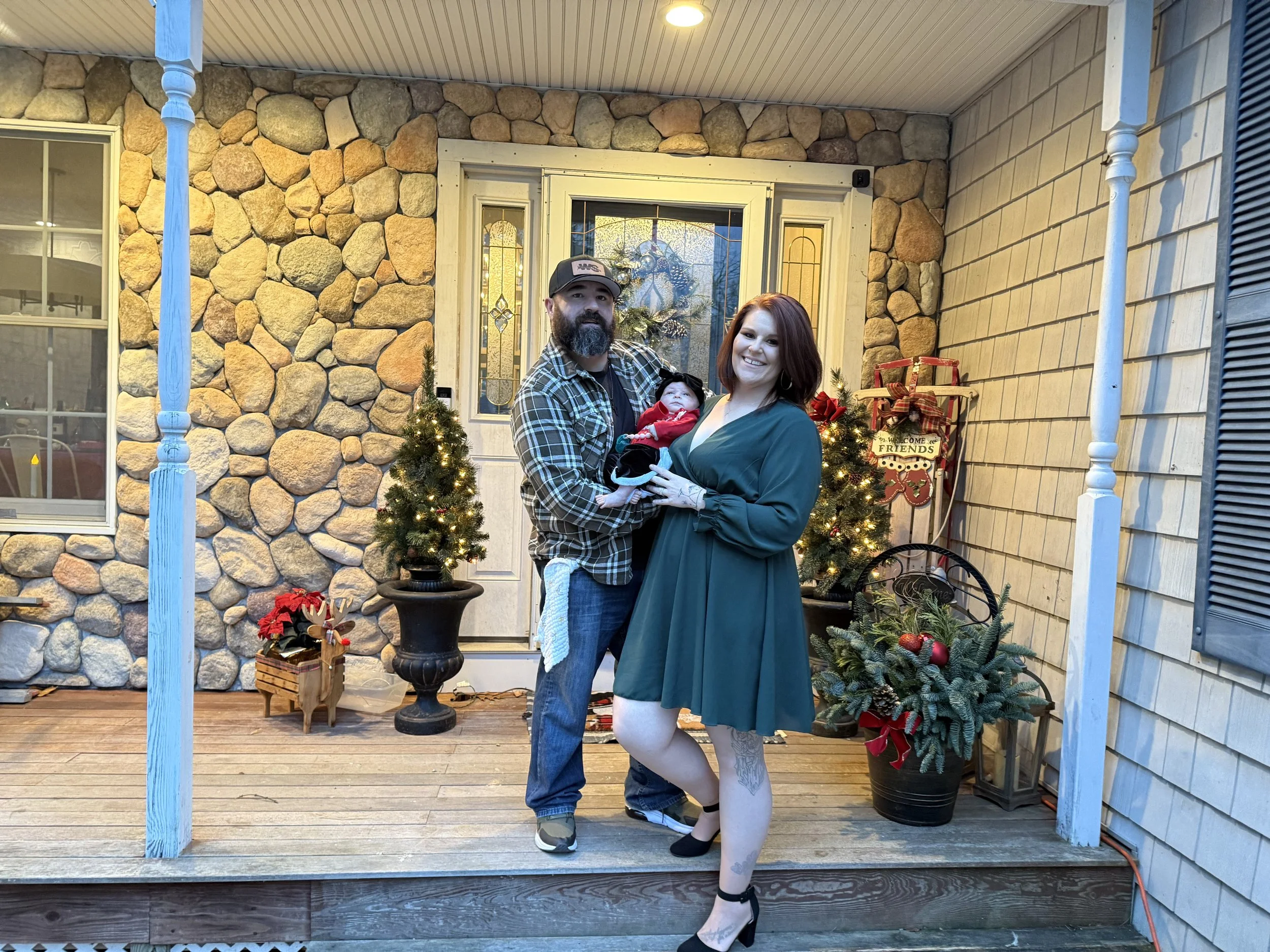 A family of three standing on a decorated porch with Christmas ornaments. The man has a beard, wears a plaid shirt and baseball cap, holding a baby. The woman in a green dress with tattoos on her legs is smiling.