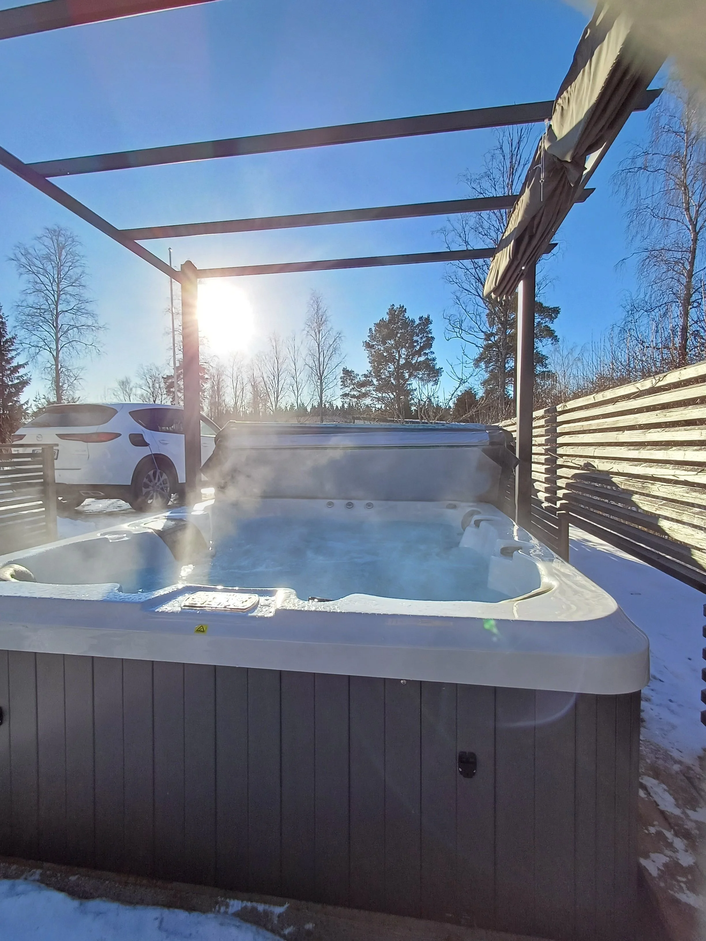 Outdoor hot tub with steam rising, surrounded by snow, with a wooden privacy fence, a wooden pergola frame, a white SUV parked nearby, and a clear sky with sunlight.