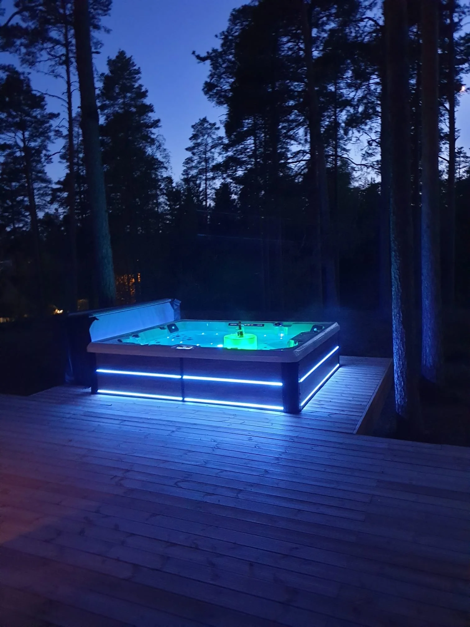 An illuminated hot tub on a wooden deck in a forest at dusk, with tall trees and a blue sky in the background.