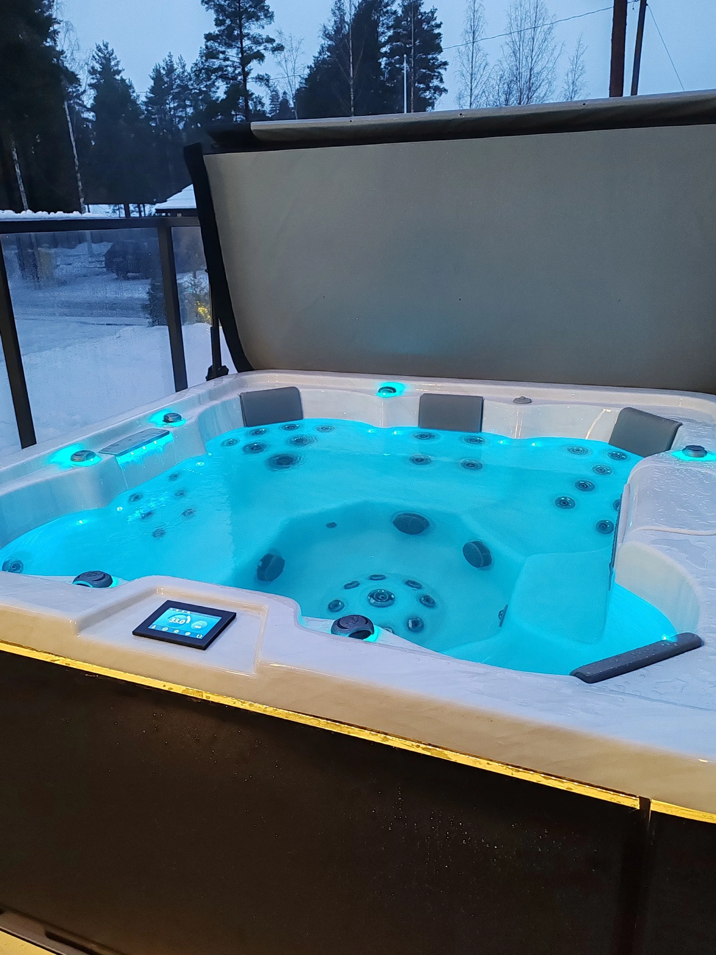 An outdoor hot tub with blue lighting, surrounded by a wooden deck, with snow and trees visible in the background.