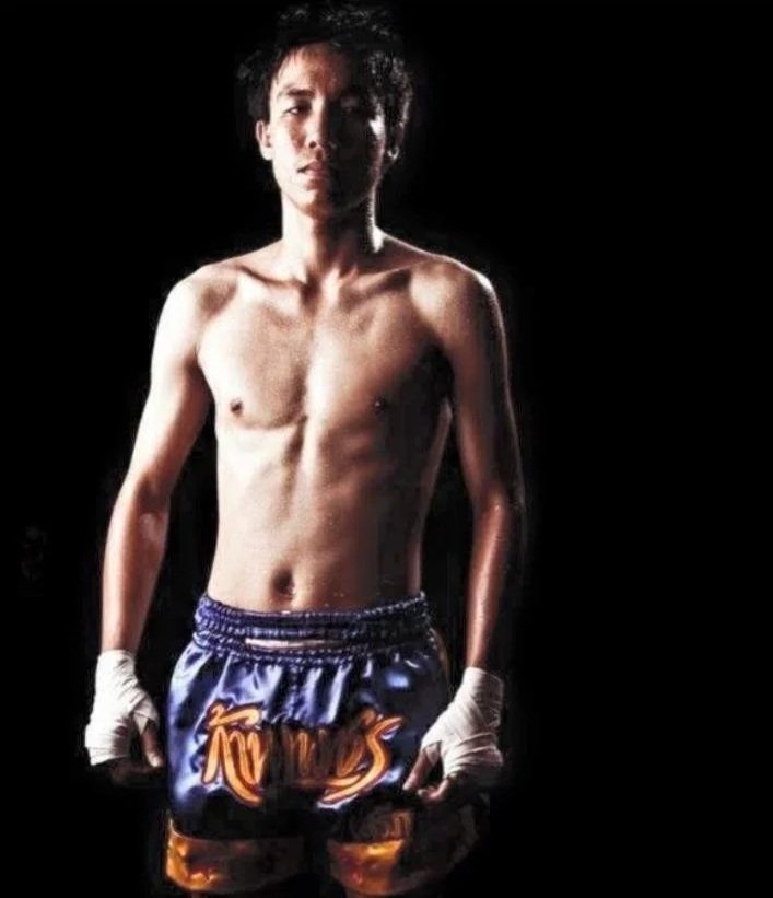 A young male boxer standing shirtless with his fists wrapped in hand wraps, wearing blue and orange boxing shorts, against a dark background.