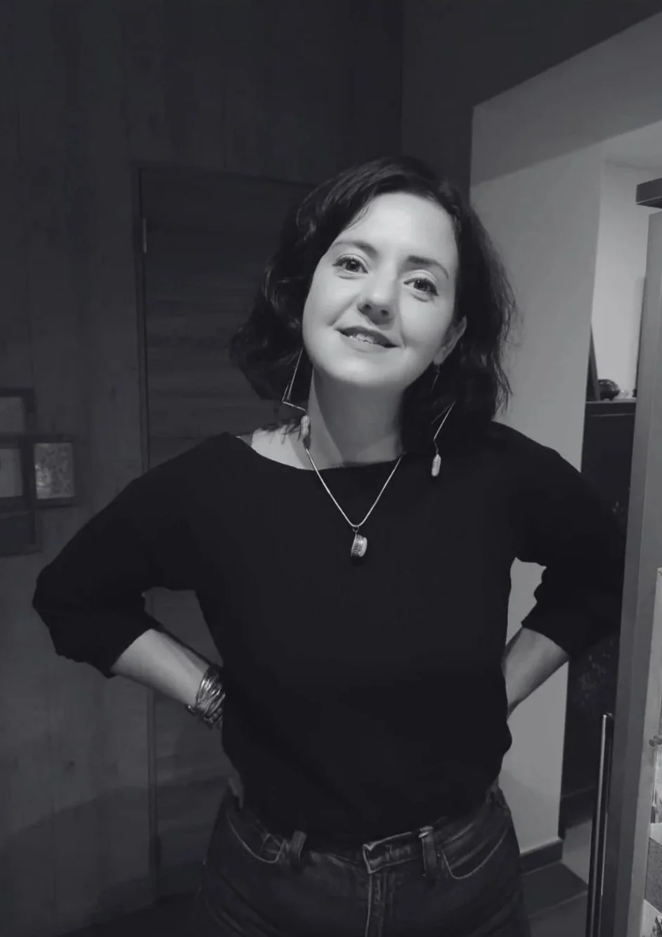 Une femme souriante avec des cheveux courts et bouclés, portant un chemisier noir, des boucles d'oreilles longues, un collier, des bracelets, posant avec les mains sur ses hanches dans une pièce intérieure.