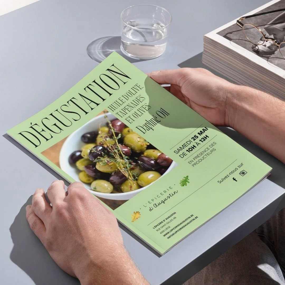 Une personne tient une affiche promotionnelle pour un événement de dégustation d'huile d'olive, avec une photo d'olives dans un bol. Sur la table devant, il y a un verre d'eau, un magazine et des lunettes.