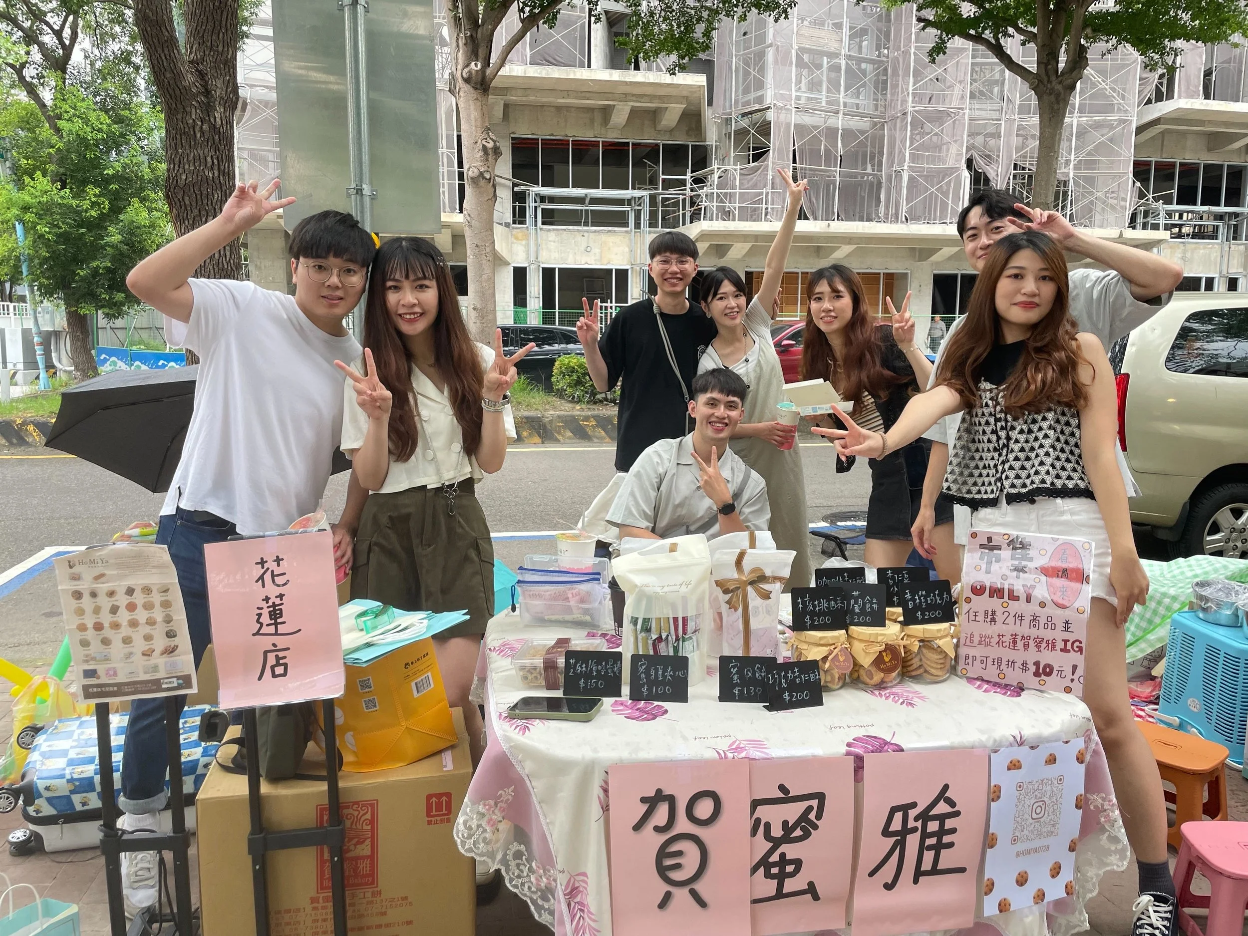 A group of eight young people posing behind a street stall decorated with pink signs and packaged baked goods, smiling and making peace signs, with a construction site and trees in the background.