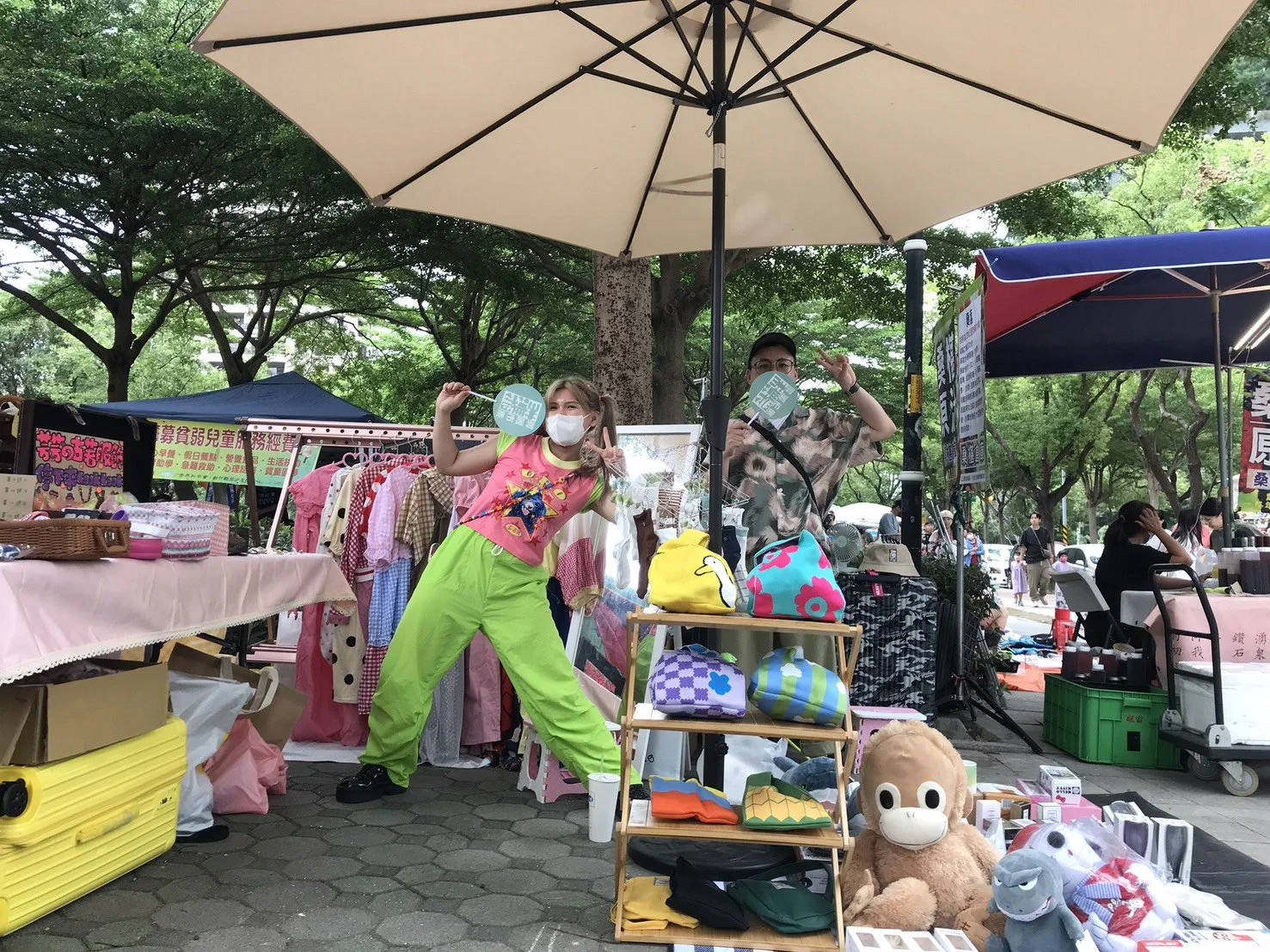 Two vendors at an outdoor market stall under a large beige umbrella, displaying colorful bags, plush toys, and clothing, with trees in the background.
