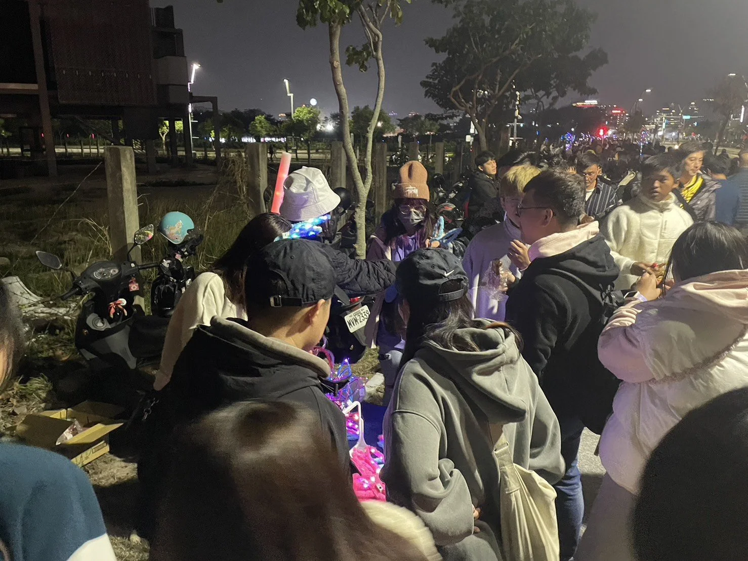 Night scene of a crowded outdoor market or street with many people gathered around various items on display, with city lights and buildings in the background.