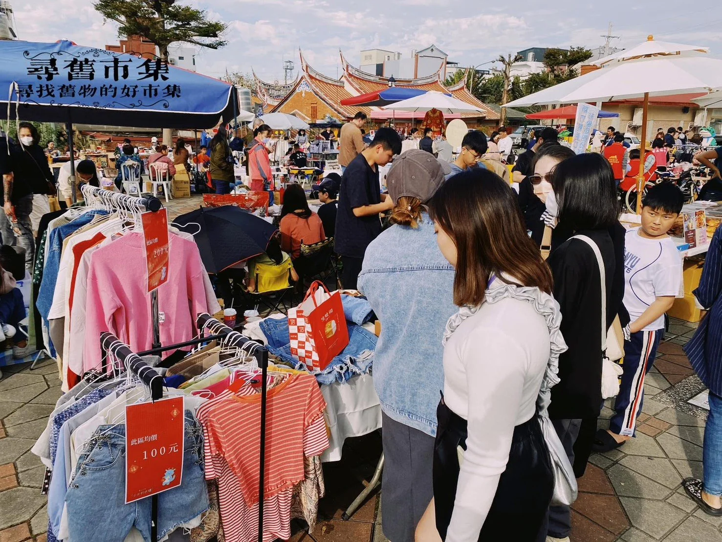 People shopping at an outdoor market with clothing and accessories, tents, umbrellas, and traditional architecture in the background.