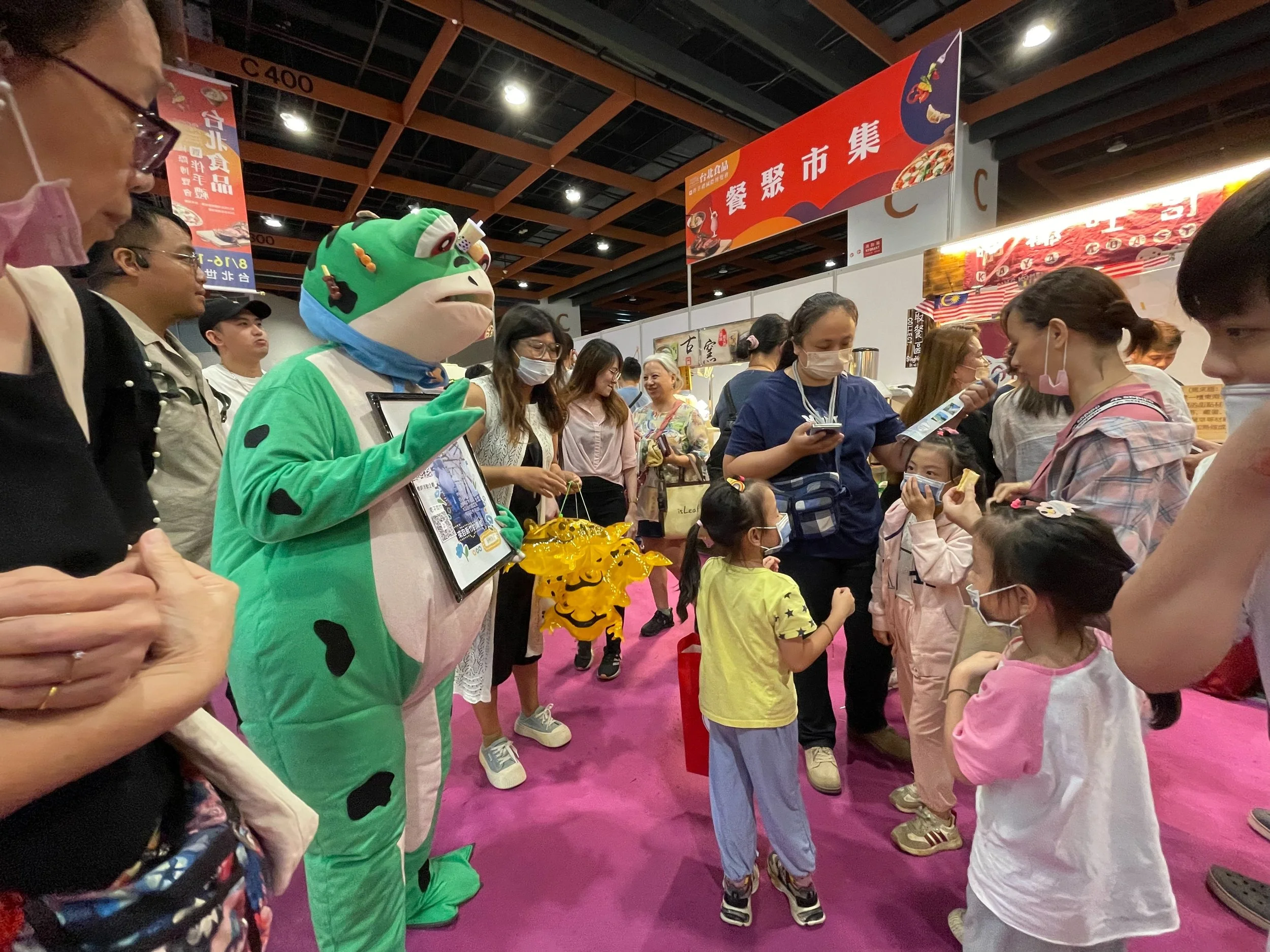 A group of children and adults are gather around a person dressed in a green animal costume at an indoor event. The person in the costume is holding a framed picture and several yellow dragon-shaped items. Some people are taking photos or looking at 