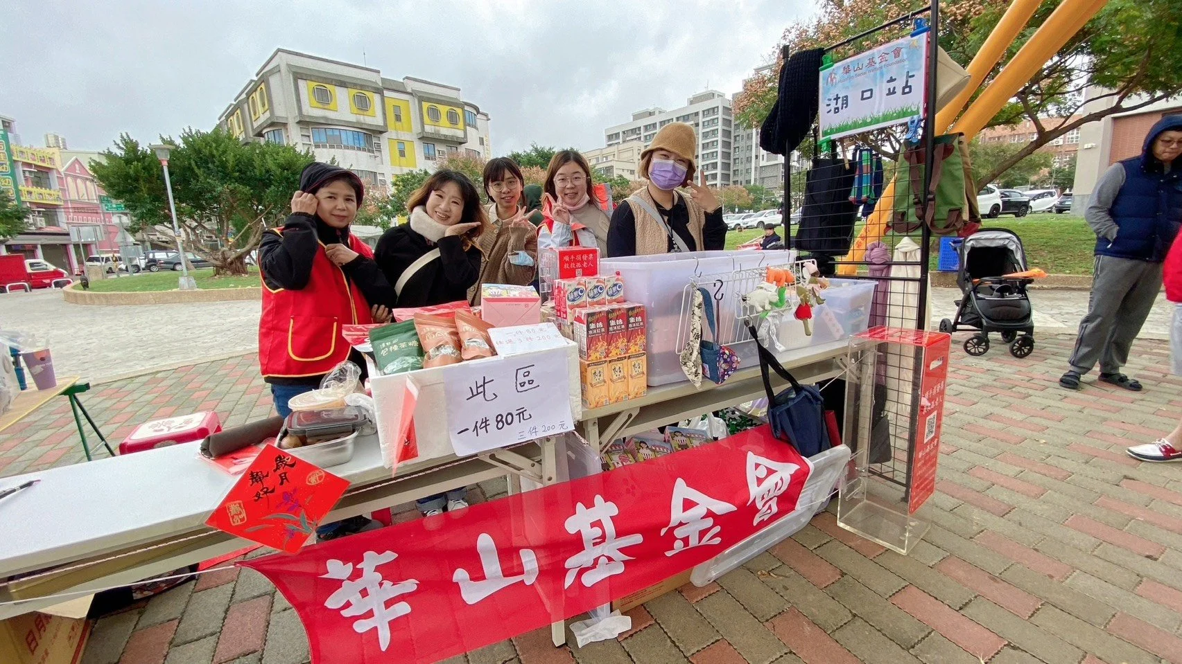 Group of people at a market stall outdoors, selling various food and goods, with buildings and trees in the background.