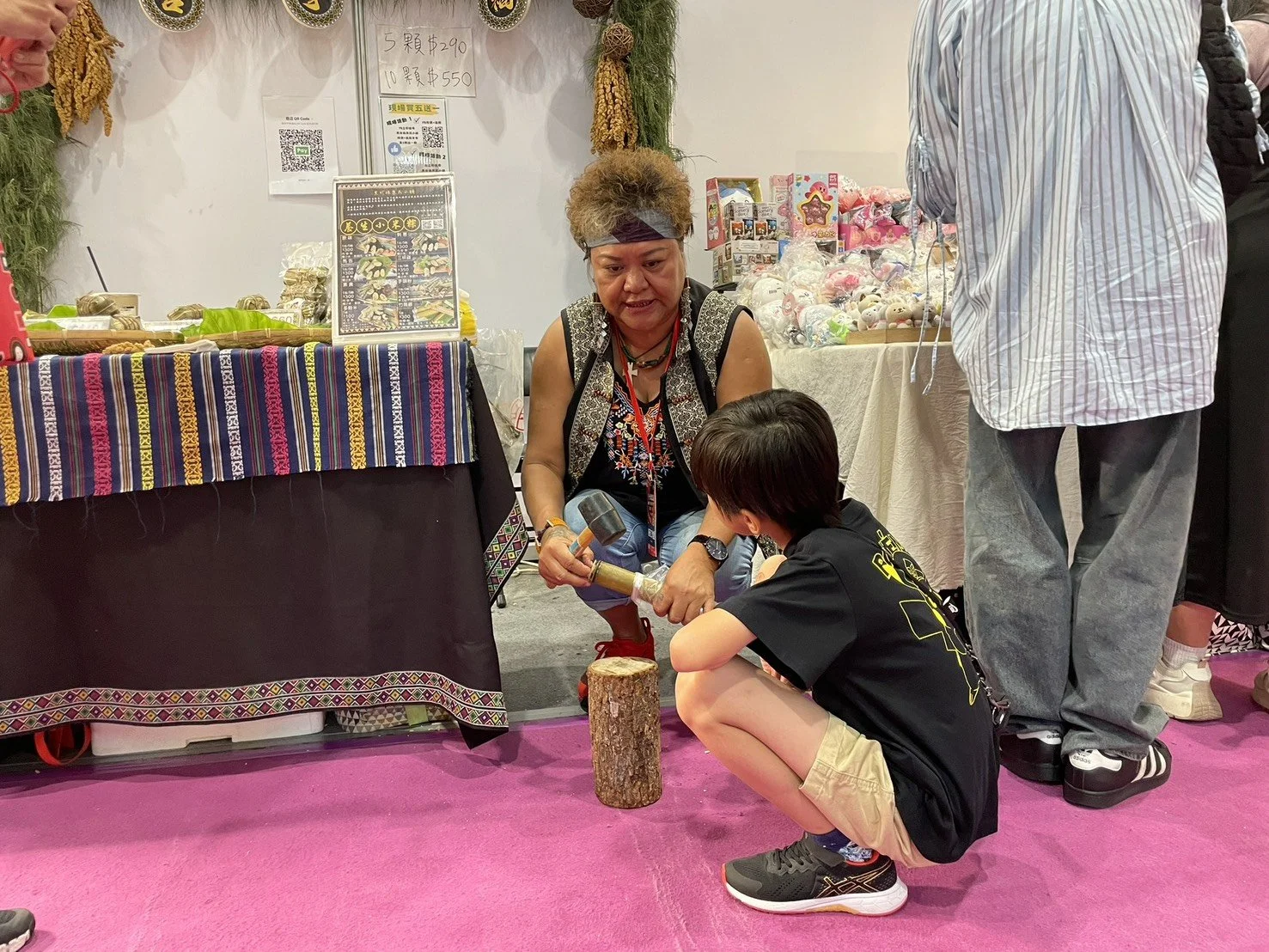 A woman with curly hair, wearing a headband and a black vest, is demonstrating how to split a piece of wood with a hammer and chisel to a young boy who is squatting in front of her. The scene takes place at a booth with colorful fabric and various it