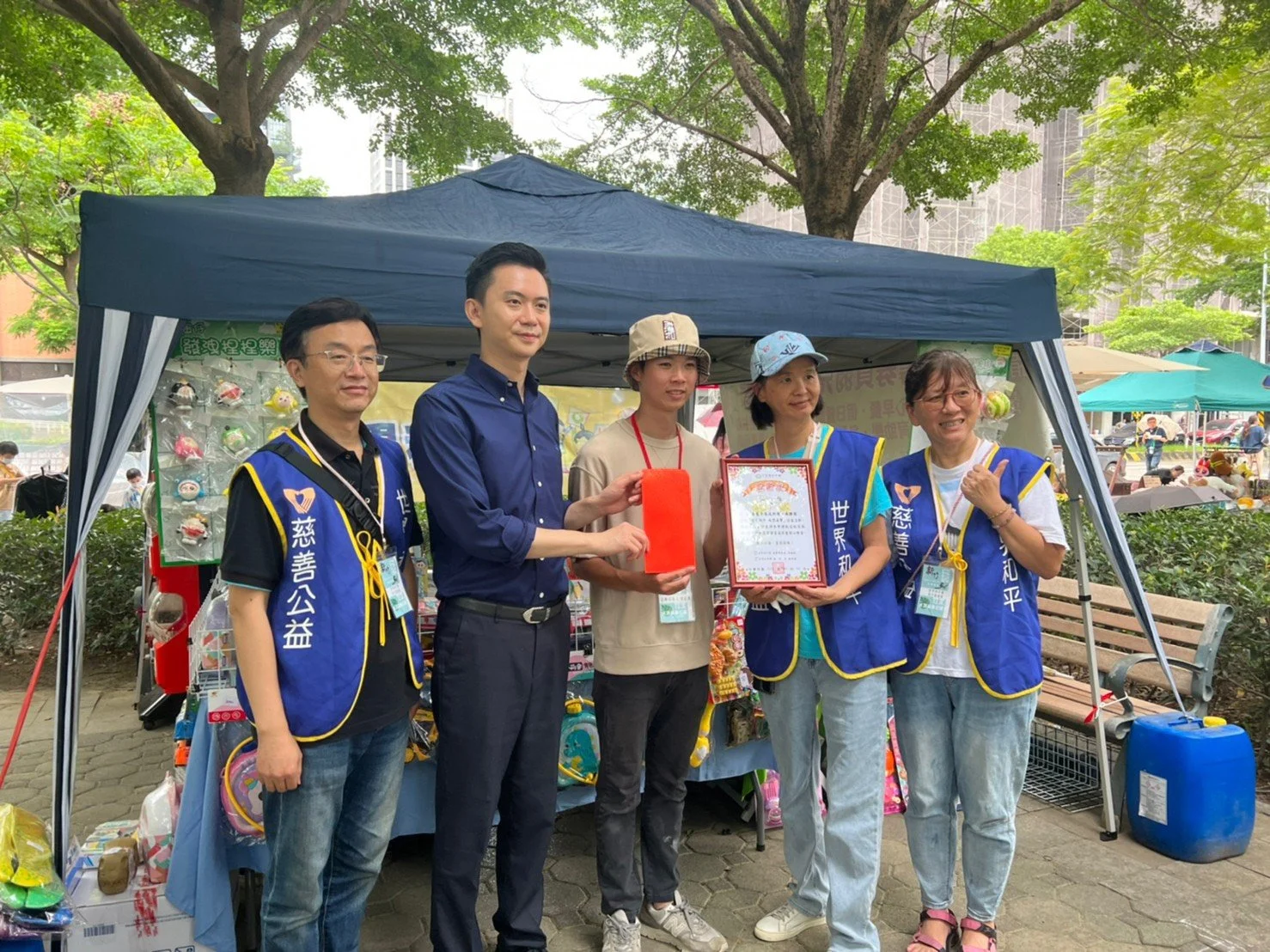 Five people standing in front of a blue canopy tent outdoors, some wearing blue volunteer vests, holding certificates and gift bags, with various items displayed inside the tent, in a park with trees and other market stalls in the background.