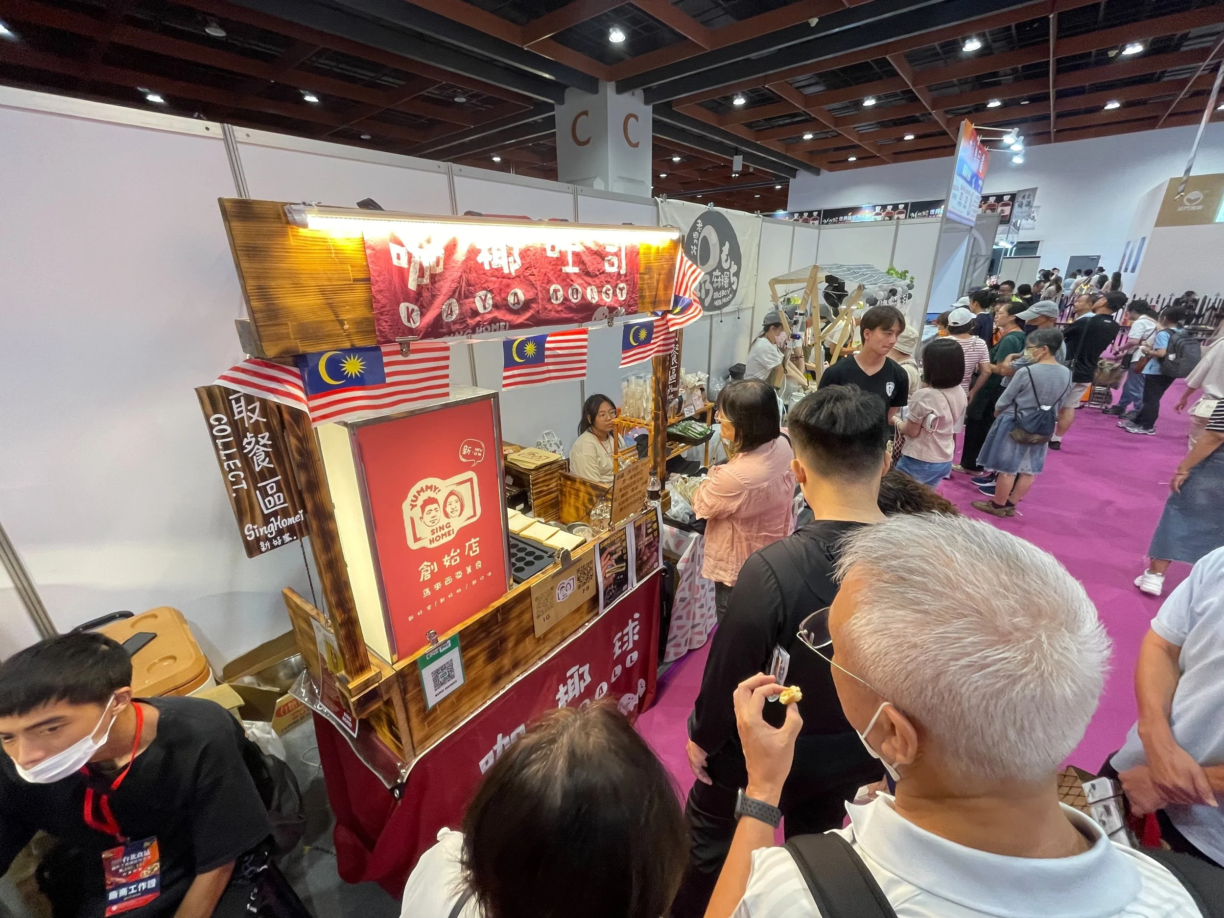 Crowd of people visiting a Malaysian food stall at an indoor festival or market, with flags of Malaysia displayed on the stall.