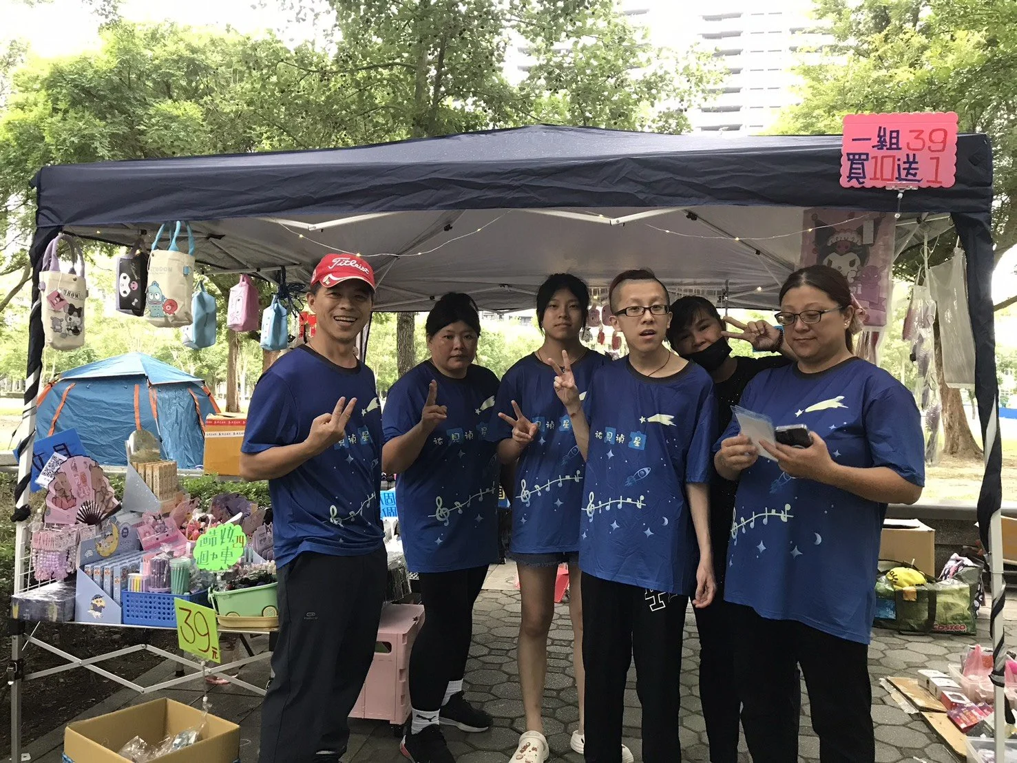 Group of six people standing together under a tent at an outdoor market, wearing matching blue shirts with space-themed designs, some making peace signs, with various items displayed on tables nearby.