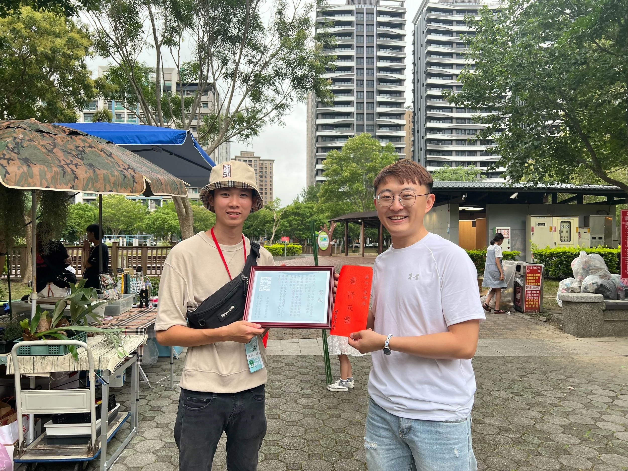 Two young men stand outdoors in a park, smiling and holding red certificates, one with glasses and the other wearing a bucket hat, with market stalls and tall buildings in the background.