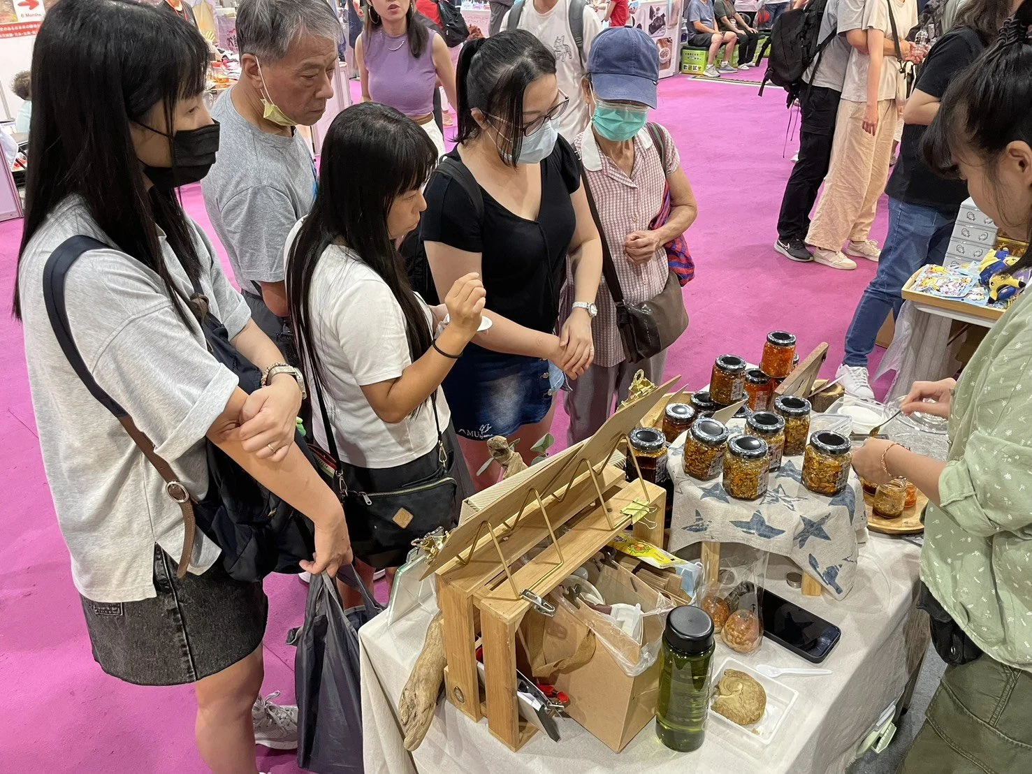 People gathered around a table at a market or fair, with jars of jam or preserves, a jar of green tea, and small food items, while an assistant is serving or selling the products.