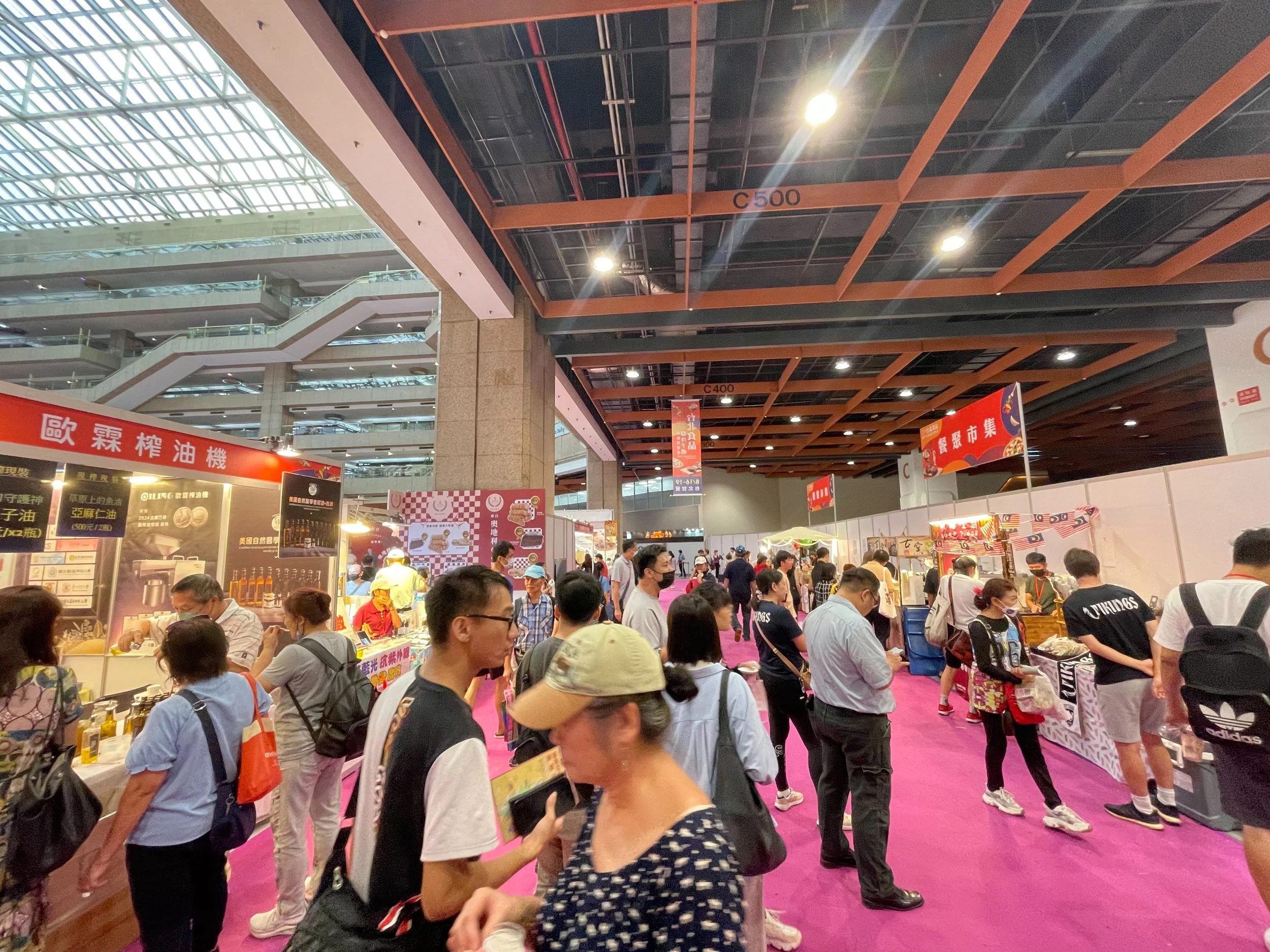 Indoor market or trade show with vendors and visitors, some wearing face masks, colorful booths, pink carpeted floor, upper level with glass ceiling, and signs in Chinese.