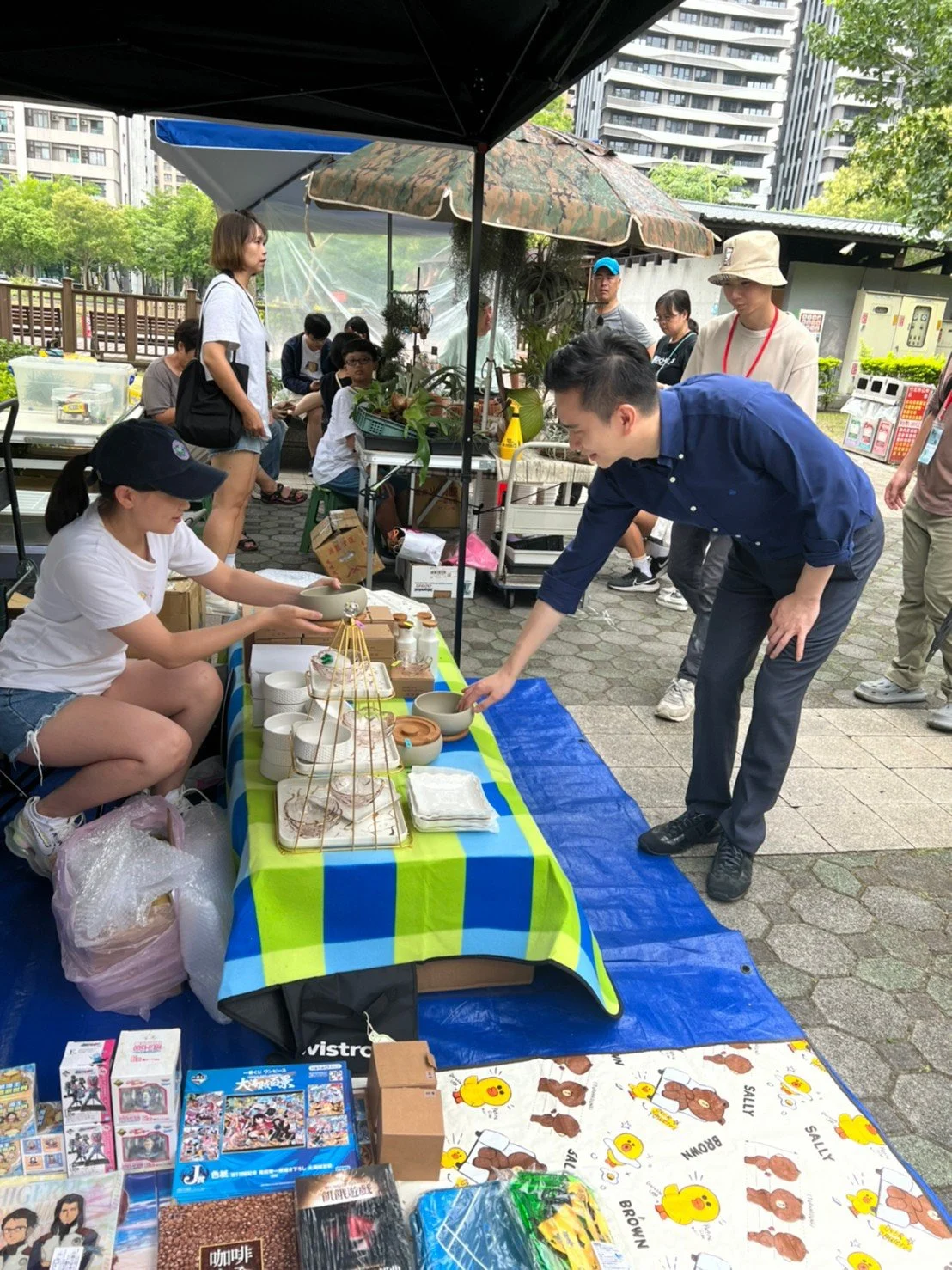 A man shopping at an outdoor market stall with ceramic bowls while a young woman, sitting behind the stall, offers items. The stall is under umbrellas with other market stalls and people in the background.