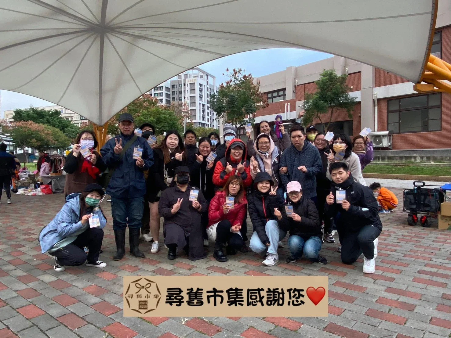 A group of people posing for a photo at an outdoor market or fair, some wearing masks and holding tickets, under a large white canopy with buildings and trees in the background, and a text banner with Chinese characters and a red heart.