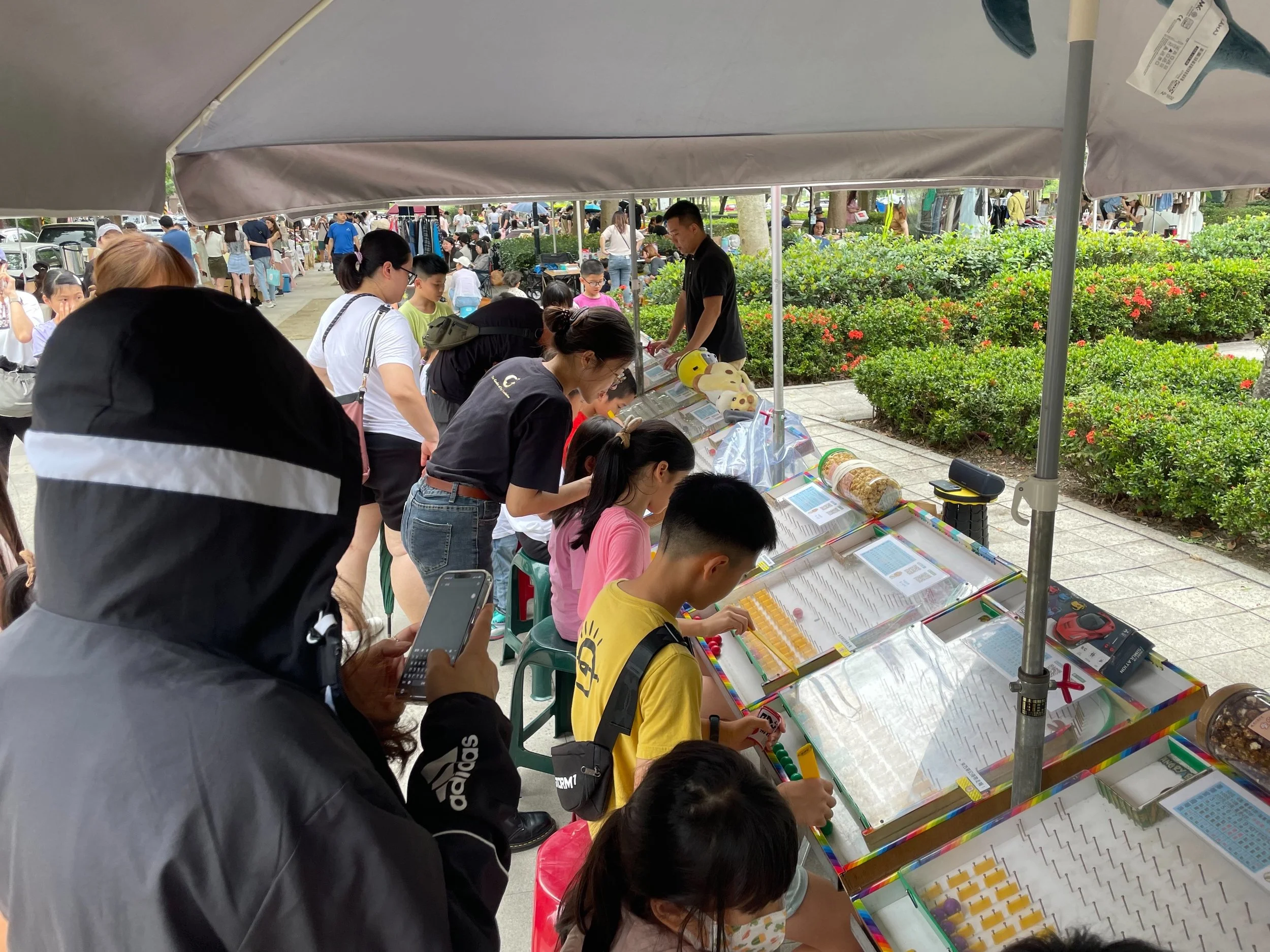 People observing exhibits at an outdoor science or educational event under a gray canopy, with plants and other attendees in the background.