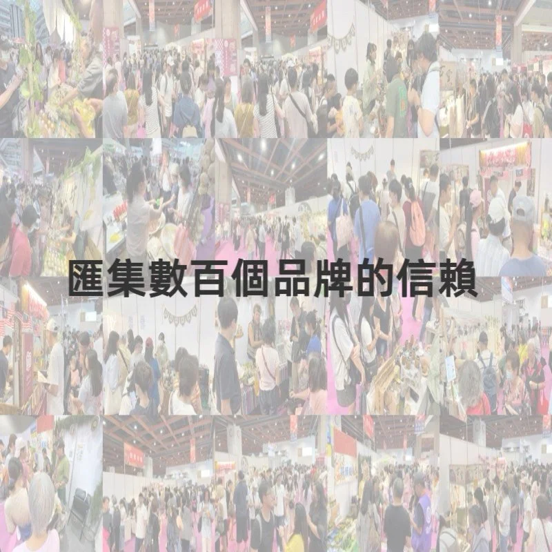 A crowded indoor market or expo with many people browsing booths and displays, some with flowers and products. The scene is lively with numerous attendees and vendor stalls.