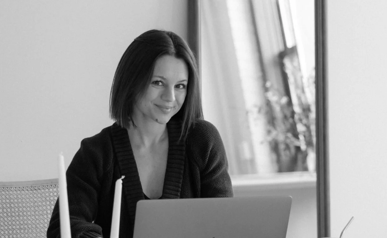 Black and white photo of Ewelina sitting behind her Macbook at a desk looking at the camera and smiling